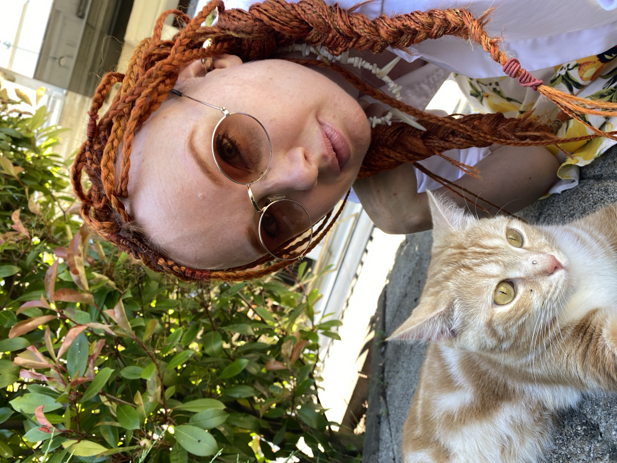 This is a close-up, upside-down, outdoor photograph featuring a person and a cat. The person, who appears to be fair-skinned with prominent freckles, has elaborate reddish-brown braided hair. They are wearing round, metallic-rimmed glasses. The cat is an orange tabby with striking yellow-green eyes and is lying down next to the person.
The scene is outdoors, with lush green foliage visible in the background. The lighting suggests it is daytime, possibly late morning or early afternoon, with sunlight filtering through the leaves. The ground appears to be a rough, grey surface, possibly concrete or stone. The overall mood is casual and relaxed, with both subjects looking towards the camera.
There is no visible text in the image. The context provided, Sochi, Russia, does not offer specific visual cues within the image itself, but it sets a geographical location for the scene.