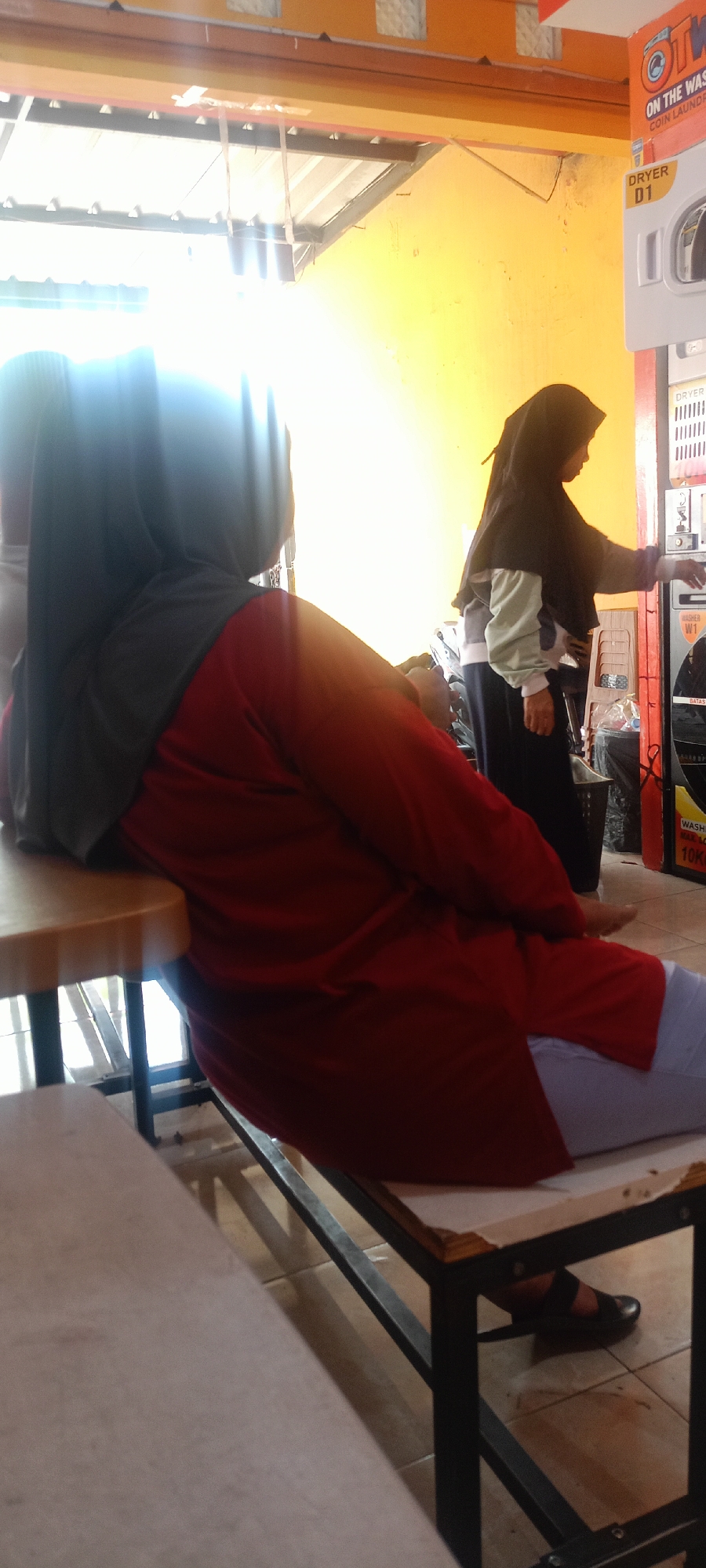 In a brightly lit indoor commercial laundry facility in Mekar Rahayu, Indonesia, two individuals are present. In the foreground, a person wearing a red tunic and a dark hijab is seated, leaning back on a light-colored table supported by a black metal frame. Their legs are crossed, and they are wearing light-colored pants and black shoes. To the right, another person wearing a black hijab, a grey and white long-sleeved shirt, and dark pants stands near a row of commercial washing machines. They are interacting with one of the machines, indicated by their hand reaching towards a control panel. The background is dominated by a bright yellow wall and overhead fluorescent lighting. The floor is tiled in a light brown color. Several washing and drying machines are visible, some with labels indicating "DRYER D1" and "WASHER W1."