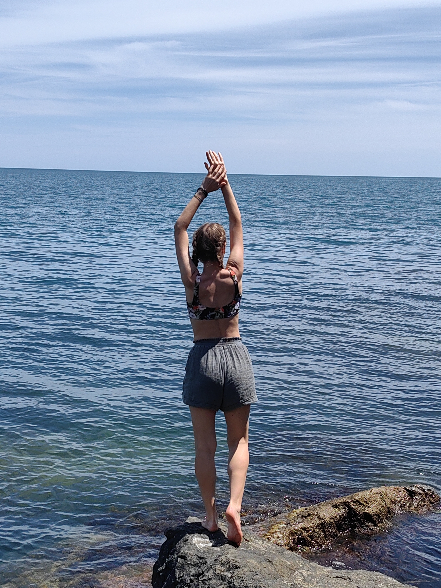 A full-body shot shows a person standing on a rock by the sea, with their back to the camera. The person is wearing a floral-patterned bikini top and grey shorts. Their arms are raised above their head with their hands clasped together. The sea is a vibrant blue, with small waves rippling across the surface. The sky above is a clear, bright blue with a few wispy white clouds. The sun is shining, suggesting it is daytime, likely midday or afternoon, and the weather is pleasant. The person appears to be stretching or posing, enjoying the natural scenery. The setting is a rocky coastline, with the water gently lapping against the shore. There is no visible text in the image.