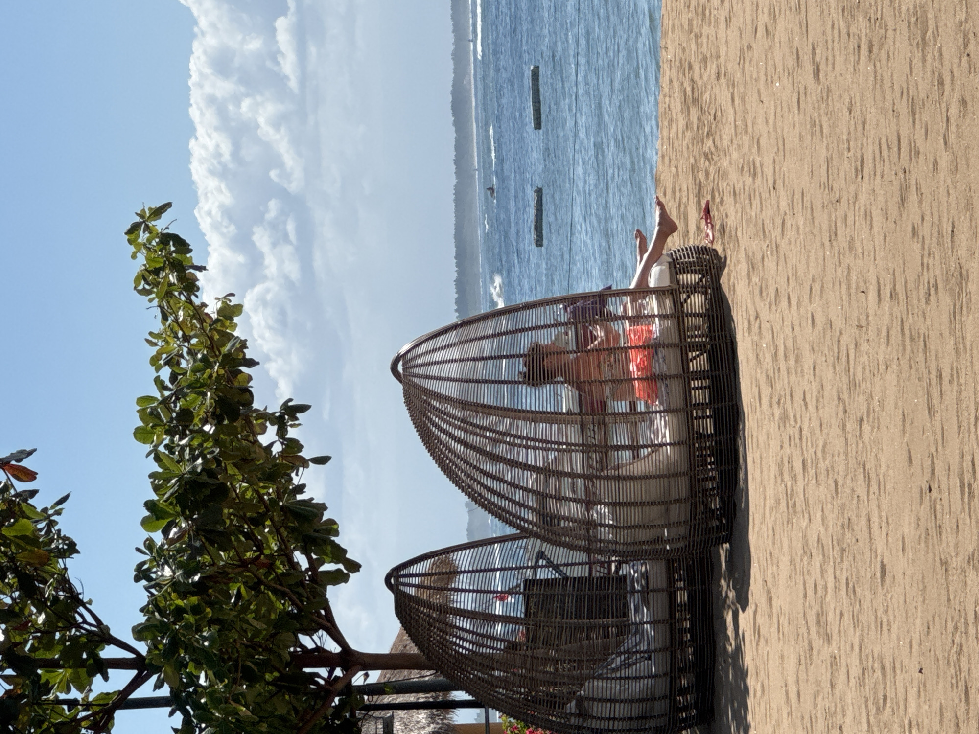 A beach scene in Kuta Selatan, Indonesia, is captured from a slightly elevated perspective. The composition includes two egg-shaped wicker chairs, one of which is occupied by a person reclining with their feet visible. A tree with green leaves frames the left side of the image. In the background, the ocean stretches towards the horizon under a sky with scattered clouds. The sand extends across the right side of the frame.