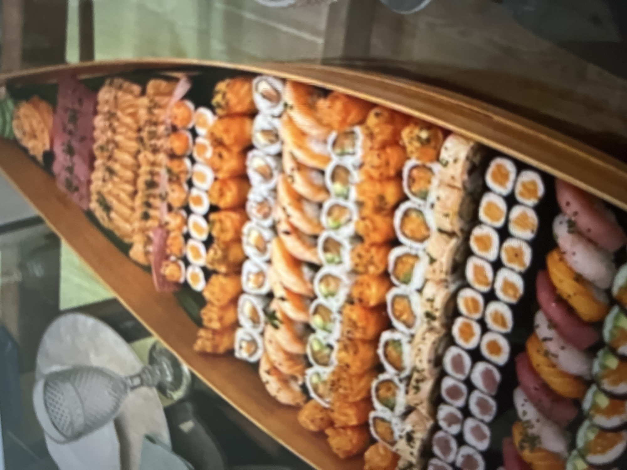 A large sushi boat is filled with various types of sushi and sashimi. The boat itself is made of light-colored wood and has a canoe-like shape. The sushi is arranged in multiple rows, showcasing a variety of colors and ingredients. Some of the visible items include nigiri with salmon and tuna, maki rolls with avocado and other fillings, and what appears to be spicy tuna or salmon on top of some pieces. The sushi is placed on dark green leaves, possibly banana leaves, as a decorative base. In the background, a clear glass with a patterned design sits on a white placemat, and a light-colored napkin is partially visible. The overall scene suggests a celebratory meal or a large gathering. The location is not specified.