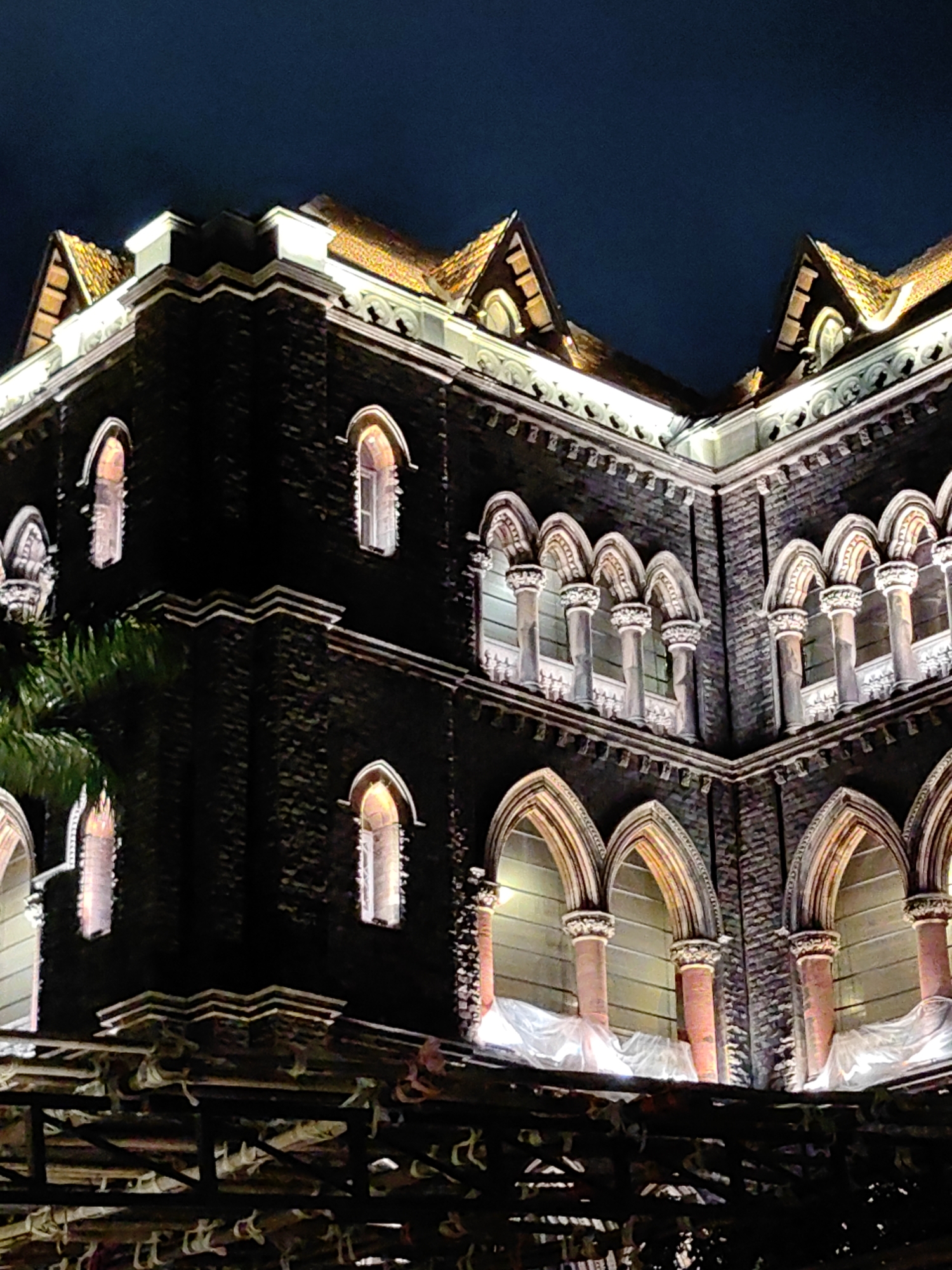 The image depicts a section of the historic Bombay High Court building in Mumbai, India, at night. The building, constructed with dark brickwork, features Gothic Revival architecture. The scene is illuminated by artificial lights, highlighting the intricate details of the facade, including arched windows with ornate stonework and pointed roofs with terracotta tiles. Several floors of the building are visible, with rows of arched windows supported by columns. In the foreground, scaffolding and some construction materials are present, suggesting ongoing work. A portion of a palm tree is visible on the left side of the image. The sky above is dark, indicating nighttime.