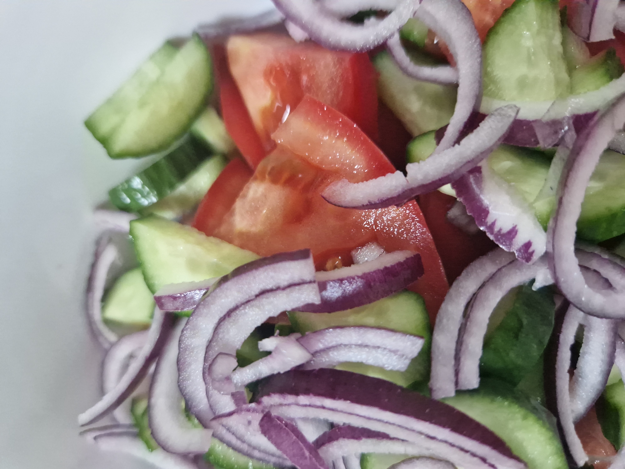 This is a close-up, overhead shot of a freshly prepared salad. The salad consists of sliced red tomatoes, sliced cucumbers, and thinly sliced red onions. The ingredients are mixed together in a white bowl, though only the contents of the bowl are visible. The lighting is bright and appears to be natural, possibly from a window. The background is a plain white surface, also likely the bowl or a table. No people or actions are depicted. The image is a still life, focusing solely on the food. The location in Sofia, Bulgaria, cannot be determined from the image itself.