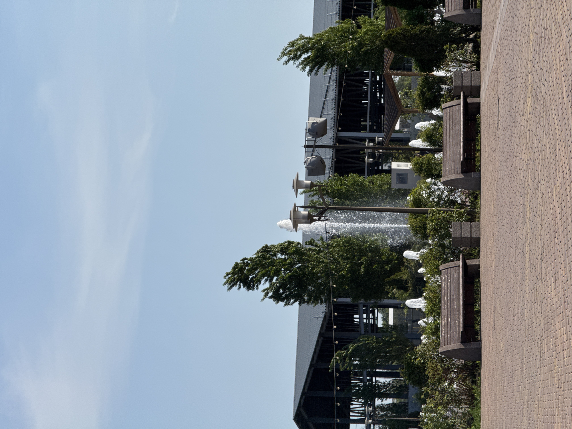 The image depicts a street scene in Rostov-on-Don, Russia. On the right is a paved walkway alongside an iron structure, possibly part of a stadium or public building. Several fountains and benches are situated along the walkway, interspersed with greenery. The sky is predominantly clear with faint wisps of clouds. The overall impression is a tranquil public space adjacent to a substantial architectural structure.