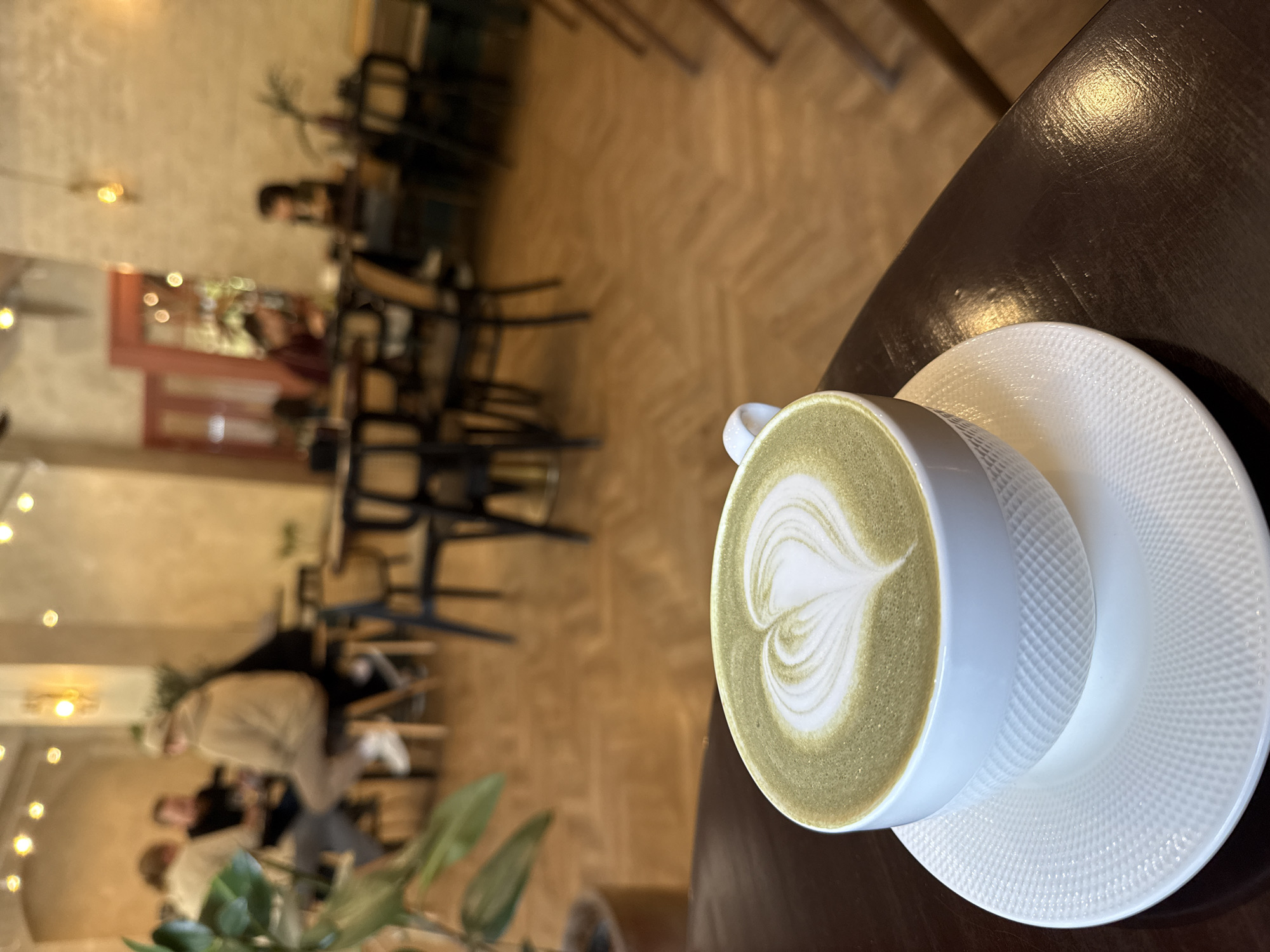 A high-angle shot shows a cup of matcha latte on a textured white saucer, sitting on a dark, possibly wooden, table in what appears to be a cafe or restaurant setting. The latte has a latte art design of a heart or similar shape on its surface.
In the blurred background, there are multiple tables and chairs, some occupied by people. The lighting suggests an indoor environment with warm, ambient light. A potted plant is partially visible in the lower left corner. The location appears to be an urban setting. It is mentioned in the request that the image is from Krasnoyarsk, Russia.