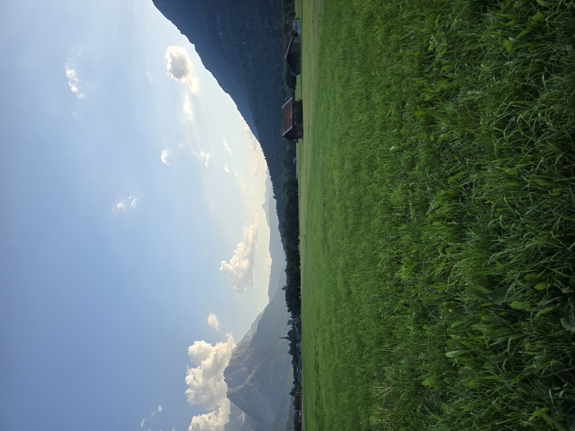The image depicts a pastoral scene with a lush green meadow stretching diagonally from the bottom right to the middle left of the frame. The meadow is bordered on the left by a shadowed mountain range, with the upper slopes covered in dark green foliage, suggesting a forested area. The sky above is a clear blue with scattered white clouds.
In the mid-ground, nestled at the base of the mountains and along the edge of the meadow, are several small buildings with dark roofs, possibly rustic cabins or barns. One building in particular, with a prominent red roof, stands out. Further in the distance, a small village or hamlet is visible, characterized by a steeple of a church piercing the horizon. The overall setting evokes a tranquil countryside landscape.
The lighting suggests it is daytime, likely late afternoon given the angle of the light casting shadows on the mountain. The weather appears clear and pleasant. The scene is serene and undisturbed, with no people or animals actively engaged in any activity. The visual cues of the mountains, the architectural style of the buildings (which are partially visible), and the general landscape strongly align with the provided location context of Garmisch-Partenkirchen, Germany. No visible text is present in the image.