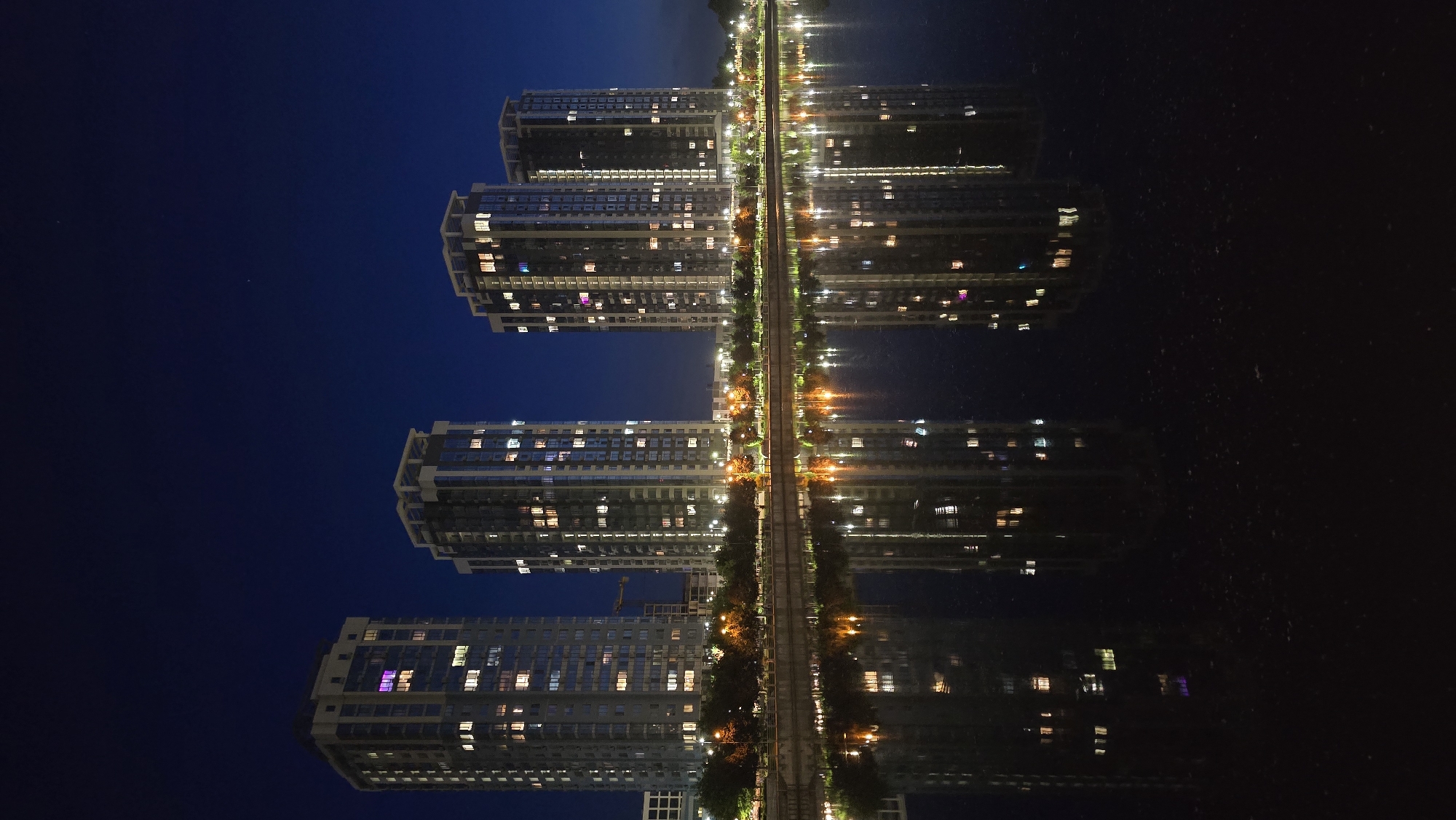 This nightscape image captures a modern urban scene dominated by high-rise buildings and their reflections. On the left side of the image, several tall apartment or office buildings stand illuminated against a deep blue night sky. Many windows in these buildings are lit with various colors, including white, yellow, blue, and touches of pink/purple, suggesting active occupancy.
A pathway or road runs vertically down the center of the image, separating the buildings from a body of water. This pathway is brightly lit by numerous streetlights and appears to have railway tracks or tram lines running along it, flanked by some trees or sparse foliage.
The right side of the image features a dark, calm body of water (likely a river or lake), which provides a near-perfect reflection of the buildings, pathway, and all the artificial lights. This creates a striking visual symmetry, with the reflections mirroring the real structures and lights across the horizontal plane of the water's surface.
The time of day is clearly night, evidenced by the dark sky and the pervasive artificial illumination. The clarity of the reflections indicates calm weather conditions with no wind disturbing the water. No human activity or visible text is discernible in the scene. The image portrays a vibrant, modern city environment at night, likely a waterfront area in Kyiv, Ukraine, given the provided context.
