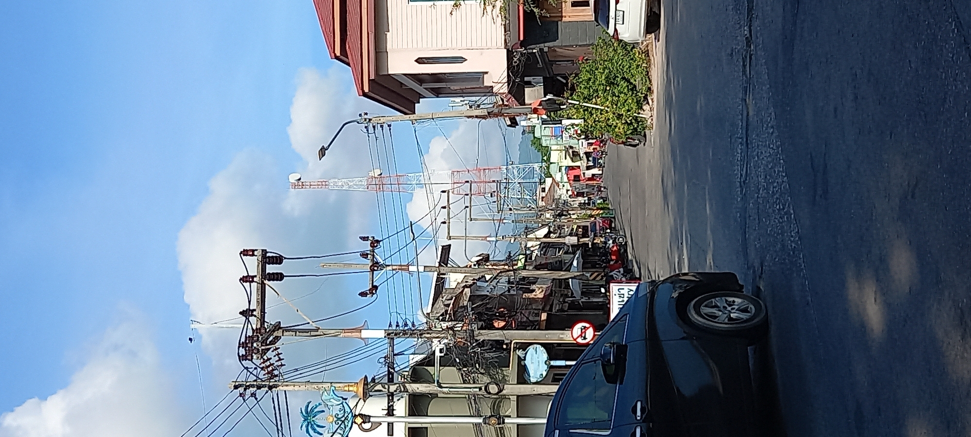 The image displays a lively street scene in a town, likely during the daytime under a bright blue sky with scattered white clouds. The perspective is slightly elevated, looking down a street lined with various buildings, some appearing to be shops or residences.
In the background, a tall red and white lattice telecommunications tower stands prominently against the sky. The mid-ground and foreground are dominated by a complex network of utility poles and wires, characteristic of urban infrastructure in many Southeast Asian countries. These poles support numerous electrical and communication lines, along with streetlights.
On the right side of the road in the foreground, a dark-colored car, possibly an SUV or minivan, is visible, either parked or moving slowly. The road surface appears to be asphalt, and further down the street, more vehicles, including motorbikes, and indistinct figures of people can be seen, suggesting a bustling environment. Green foliage and bushes punctuate the roadside.
Several signs are visible:
1. A white rectangular sign near the black car displays Thai text: "เข้า ซอย". "เข้า" (Kao) means "enter" or "in," and "ซอย" (Soi) means "alley" or "lane." Thus, it indicates an "Entrance to Lane/Alley."
2. Below this, a circular sign features a red border, white background, and a black motorcycle symbol with a red diagonal line through it, indicating "No Motorbikes" or "Motorbikes Forbidden." This could signify a restriction on motorbike entry or parking.
3. Lower on a utility pole, a decorative blue circular sign with a palm tree and waves motif also contains Thai text: "ฟ้าใส" (Fah Sai), meaning "Clear Sky" or "Bright Sky," which is a common business or place name.
The bright sunlight and clear sky suggest a sunny day, likely mid-morning or afternoon. The overall scene depicts a typical street in an urban or semi-urban area, consistent with the given location context of Thong Sala, Thailand.