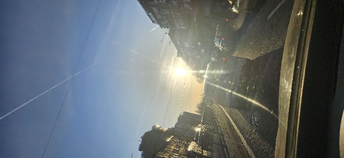 The image depicts an urban street scene in Varna, Bulgaria, viewed from the interior of a vehicle during either sunrise or sunset, indicated by a low-angle sun. The bright sun is positioned centrally in the upper mid-ground, causing significant lens flare and obscuring fine details.
The sky is clear and light blue, traversed by two prominent white contrails that intersect in the upper-left quadrant, alongside several fainter contrails. Thin, dark overhead lines or cables are visible horizontally strung across the street, silhouetted against the sky.
The foreground shows the top of a vehicle's dashboard. The street is a multi-lane asphalt thoroughfare, with numerous vehicles in what appears to be traffic. Directly ahead is the rear of a light-colored passenger car or SUV; its license plate is obscured by sun glare. Other vehicles are visible further down the road. The road surface reflects the intense sunlight.
On the left side of the street, a dark, decorative metal railing runs parallel to the road. Behind it stand multi-story buildings of urban architectural style, featuring multiple windows. Sparse, possibly bare trees or bushes are situated between these structures. On the right side, additional multi-story buildings line the street, though their specific details are less distinct due to backlighting and glare.
No individuals are clearly discernible within the frame. The scene conveys an active urban environment with vehicular traffic under bright, direct sunlight on an unidentified urban street within Varna, Bulgaria.