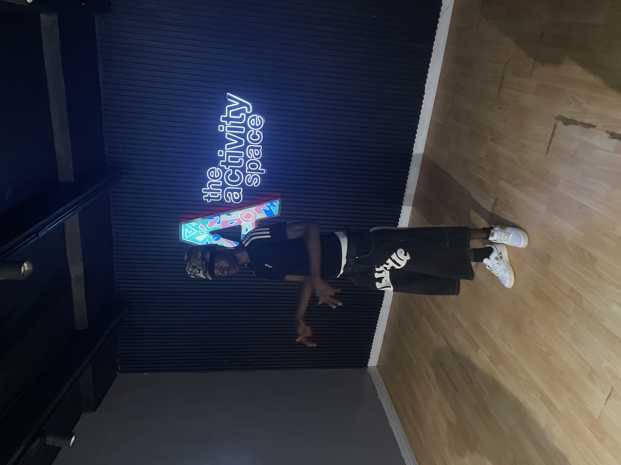 The image, rotated 90 degrees clockwise, displays an individual standing in an indoor setting. The background features a dark, vertically ridged wall, potentially composed of acoustic panels, with a bright blue neon sign. A light-colored wooden floor occupies the right side of the frame.
The main subject is a young person, centrally positioned, wearing a black t-shirt with white stripes on the sleeves, black shorts featuring white stylized text on the side, a camouflage-patterned cap, and white sneakers. They are captured in a dynamic pose, with one arm bent and hand open, looking directly towards the camera.
A prominent feature of the setting is the neon sign on the dark wall, which clearly reads "the activity space" in glowing blue letters. Below this, another colorful neon graphic with geometric patterns is visible. The wooden floor shows some signs of wear, including scuff marks and darker lines, suggesting it is a well-used area. The lighting is artificial, highlighting the neon sign and the subject, indicative of an indoor recreational or studio environment. The overall scene suggests a contemporary space, possibly a dance studio or community center in Lekki, Nigeria.