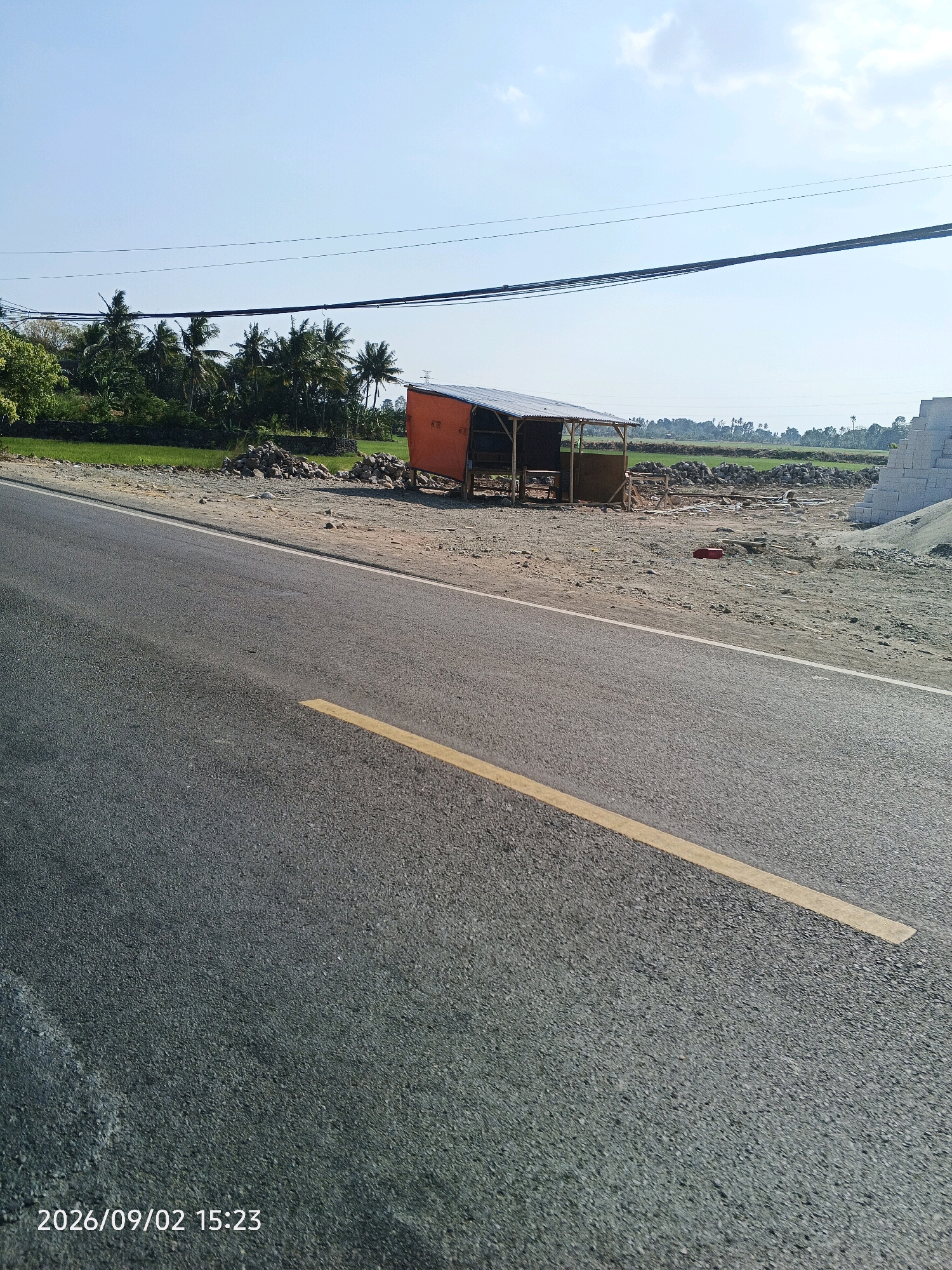 The image depicts a roadside scene in Pinrang, Indonesia, on September 2, 2026, at 15:23. A paved asphalt road with a yellow dashed line runs across the foreground, angled from the bottom left towards the center right. To the right of the road, a small, open-sided shelter with a corrugated metal roof and orange siding is visible. The shelter is supported by wooden posts and appears to be a simple structure, possibly for vendors or laborers. Adjacent to the shelter, piles of rocks and construction materials, including what appear to be concrete blocks, are scattered on the dusty ground. In the background, a lush green field stretches out, dotted with palm trees, characteristic of the tropical landscape. The sky above is clear and blue with a few wispy clouds. Overhead, dark power lines are strung across the frame. The overall scene suggests a semi-rural or developing area along a road.