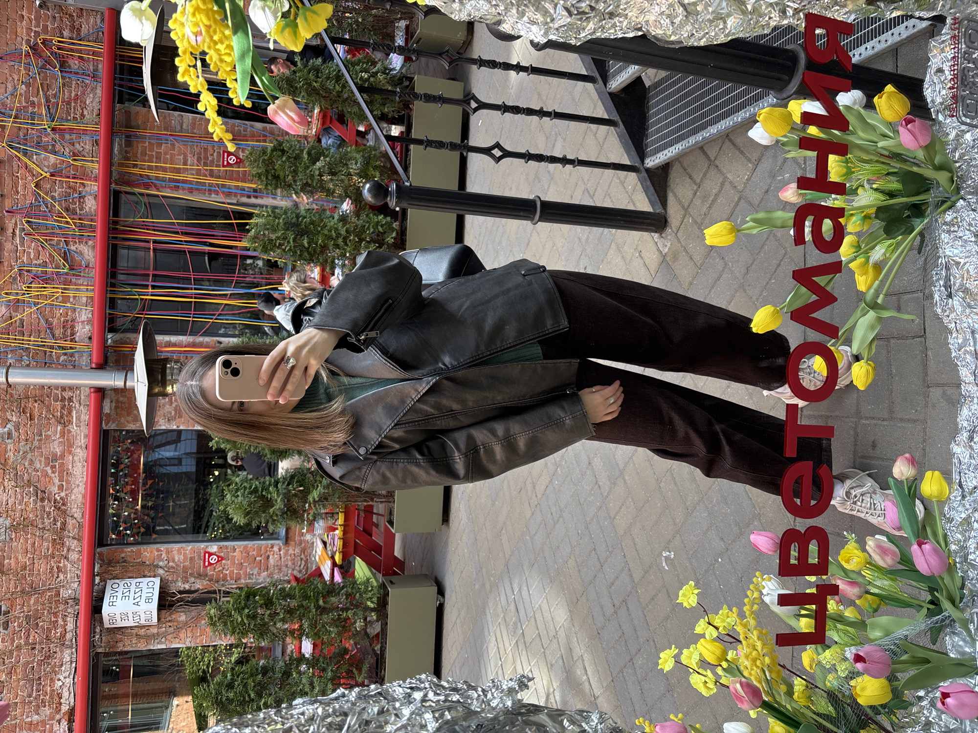 A woman is taking a selfie in front of a store displaying "цветомания" (flower mania) in Saint Petersburg, Russia. The background includes a brick building decorated with colorful wires, greenery, and decorative elements. The woman is wearing a black leather jacket and brown pants. The storefront is decorated with flowers and tinfoil. The ground is paved with gray bricks. The reflection is of a stairway with black metal railings.