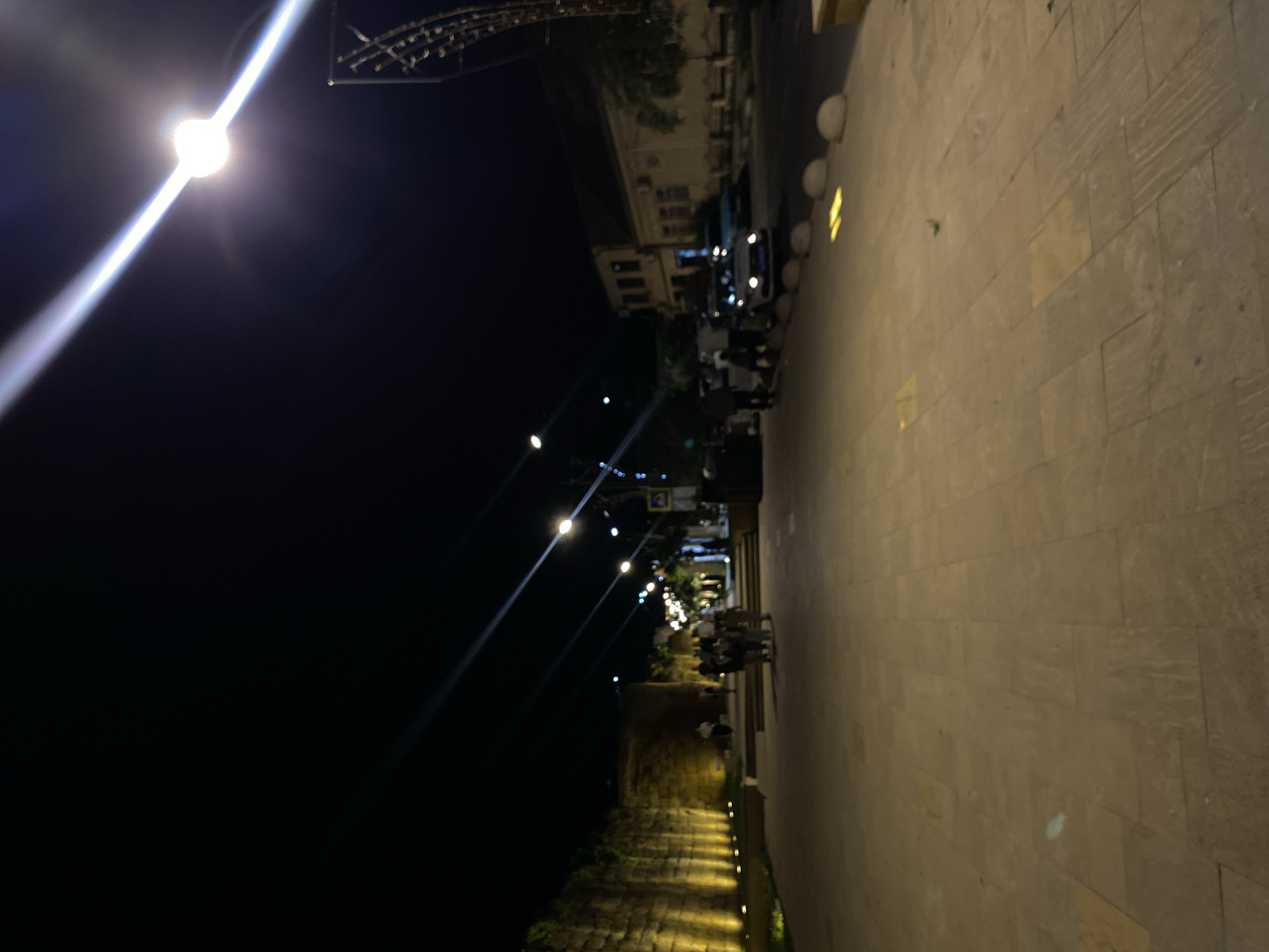 The image depicts a nighttime scene on a paved pedestrian walkway in Derbent, Russia. The walkway is illuminated by warm-toned lights along a textured stone wall on the left and by streetlights overhead, casting long shadows and lens flares across the scene. Several individuals are walking on the walkway and gathered in small groups. On the right side of the walkway, a street is visible with parked cars, including what appears to be a police car with its lights on, and buildings with illuminated windows. The overall environment suggests a public space in a city at night, likely a promenade or street in Derbent.