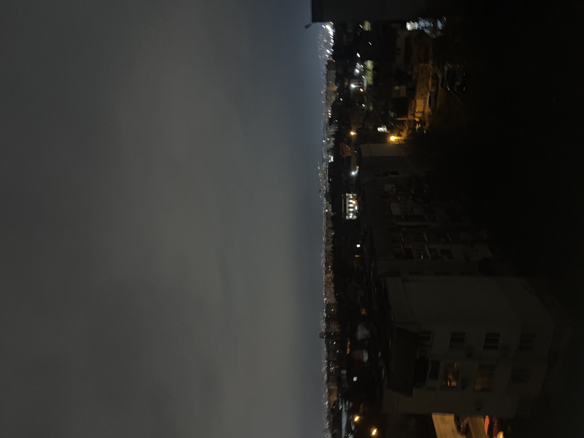 The image shows a nighttime view of Sofia, Bulgaria. On the right side of the image, there is a building with several windows visible, some of which are illuminated. Streetlights and city lights stretch out along the horizon in the center of the image, indicating the urban sprawl of Sofia. The left side of the image is dominated by a dark, overcast sky. The overall atmosphere is dark due to the night and cloud cover.