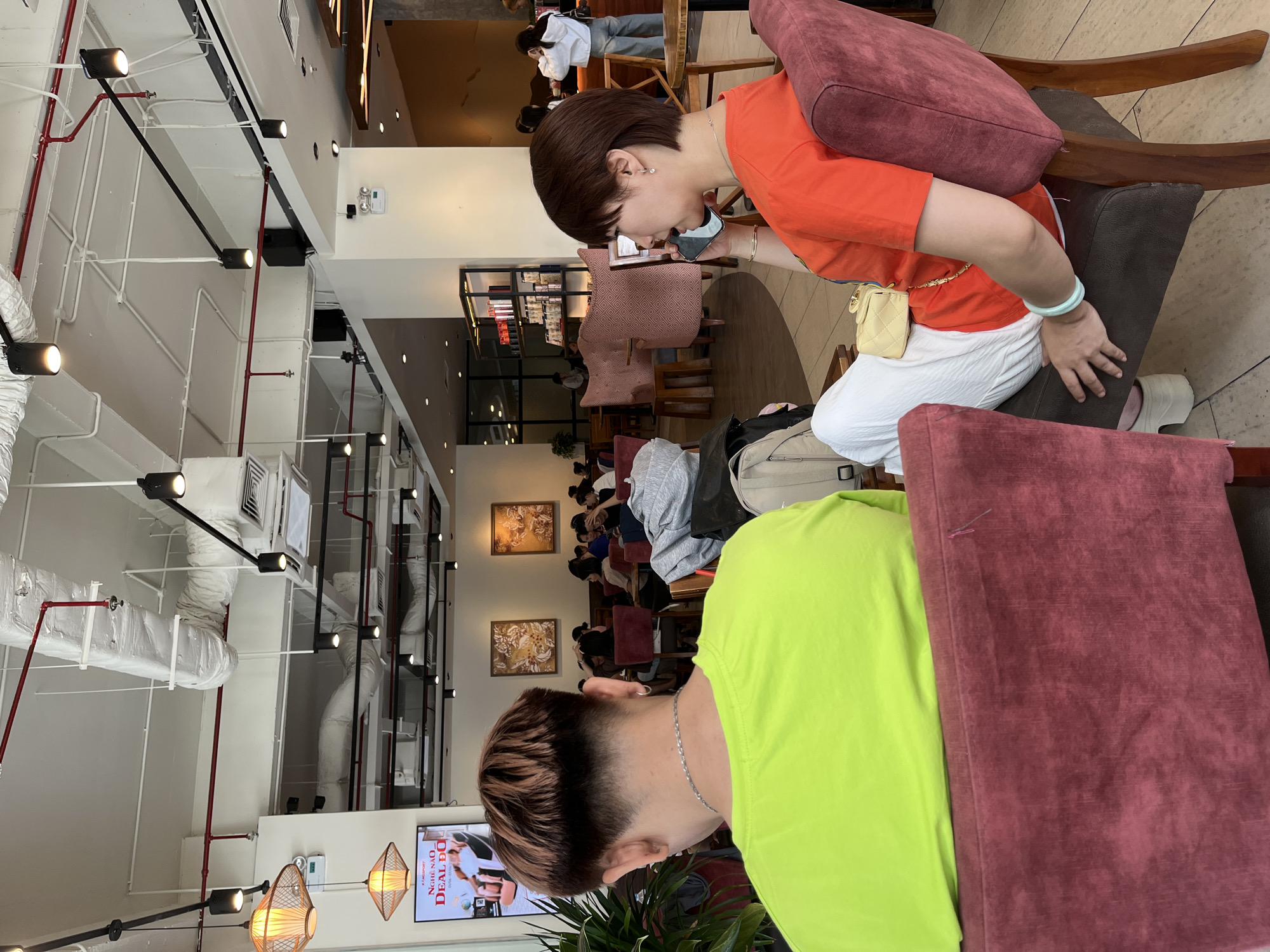 The image captures an indoor scene, likely a modern cafe or restaurant, in Đà Nẵng, Vietnam. The setting features an industrial-chic aesthetic with an exposed ceiling, revealing white and red pipes, black track lighting fixtures, and ventilation ducts.
In the immediate foreground, to the right, a person with short, dark hair, styled with lighter highlights and a faded cut, is seen from behind, wearing a bright lime green t-shirt. Seated opposite, on the left, is a woman with short, dark brown hair, wearing an orange t-shirt, white textured pants, and white clogs. She is actively engaged with her mobile phone, holding it up to her face as if on a call or using an app. Her accessories include a silver necklace, pearl earrings, and a light green bangle on her right wrist, along with a small, yellow quilted handbag worn across her body.
The background shows several other patrons seated at tables, some also looking at their phones, indicating a casual social or waiting environment. The furniture consists of chairs with dark brown wooden frames and red or patterned upholstery. Decorative elements include framed artworks on the walls and warm-toned woven pendant lights.
A television screen in the background displays text, which includes Vietnamese phrases such as "NGHĨ NÀO" (What do you think/Think of something) and "DEAL ĐÓ" (That deal), along with "x La Mer..." partially visible, confirming the location context in Vietnam. The overall atmosphere appears relaxed and brightly lit by a combination of artificial lighting.