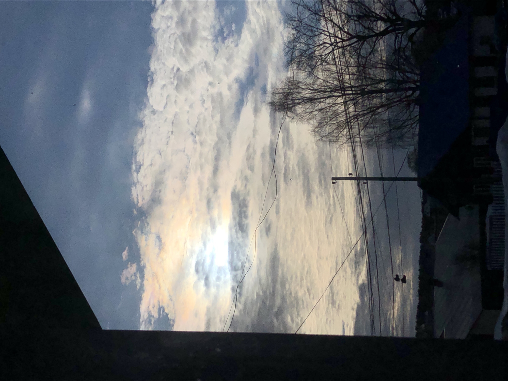 The image presents an outdoor scene captured from an elevated perspective in the city of Foki, Russia. A dark, angled architectural element occupies the left side and top-left corner of the frame, indicating the point of view. The sky is partially cloudy, featuring a mix of stratocumulus and altocumulus clouds, with visible patches of blue sky. A bright, partially obscured sun, exhibiting iridescent halo effects, is visible in the upper-left quadrant through the clouds. Multiple dark power lines or utility wires span horizontally and diagonally across the midground. A dark utility pole with crossbars stands slightly left of center, supporting some of these wires. A pair of dark shoes hangs by their laces over one of the power lines in the left-center. Several deciduous trees, bare of foliage, are prominently silhouetted in the right midground. In the background, a residential area consists of houses with dark, sloped roofs, and patches of white material, likely snow, cover the ground. A distant tree line marks the horizon. No individuals are present in the scene.