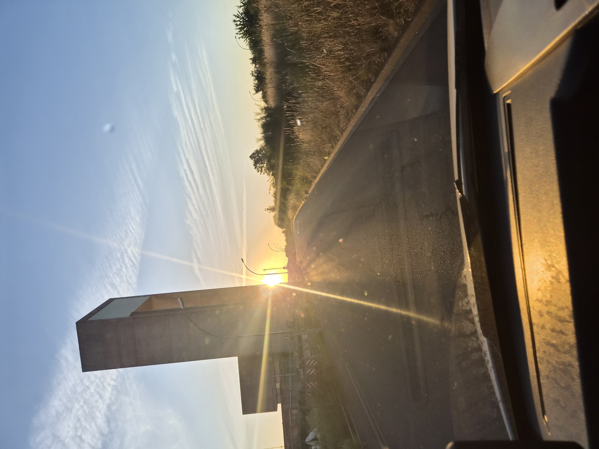The photograph depicts a view from inside a vehicle, looking out towards a road and a large concrete structure at sunset. The sun is low on the horizon, casting strong lens flares across the image and illuminating the sky with warm hues of orange and yellow. Wispy clouds are visible in the bright blue sky. The concrete structure, which appears to be a modern building or monument, is tall and angular, with a section of glass at the top. To the right of the road, there is dry, tall grass and some trees. The foreground shows a portion of the vehicle's dashboard and windshield, with the windshield being speckled with dirt and showing reflections. The road ahead curves gently. In the distance, lampposts are visible. The scene suggests a drive through an area on the outskirts of a city or town during the golden hour. Specifically, the concrete structure is identified as the "Bureaux Fédéraux" in Leudelange, Luxembourg.
