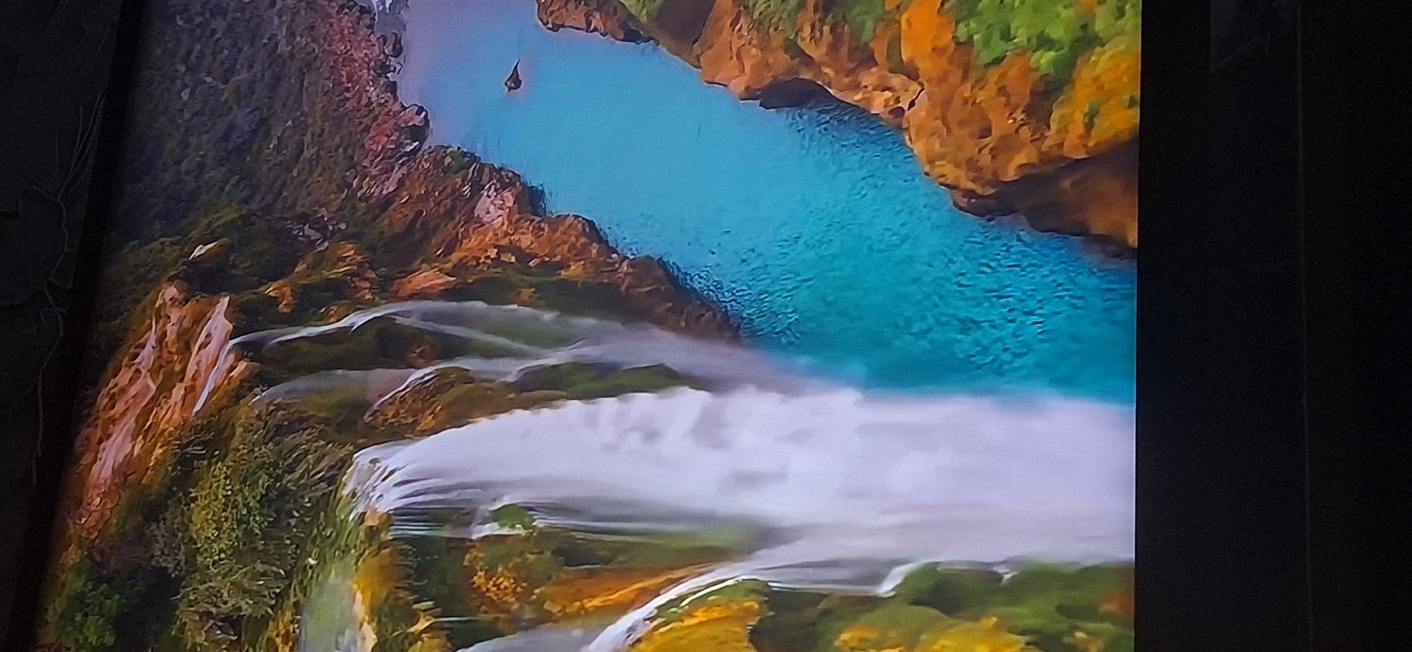 The image displays a vibrant, high-angle view of a cascading waterfall plunging into a turquoise river. The waterfall originates from a lush, green cliffside with some reddish-brown rocky outcroppings. The river flows alongside the cliff, with more rocky terrain and vegetation lining its banks. The water in the river is clear and reflects the surrounding colors. A small, dark bird is visible flying above the river to the right of the waterfall. The overall scene is one of natural beauty and tranquility. The image is captured on what appears to be a screen, with a dark border at the top and bottom, suggesting it is a photograph of a display. There is no discernible connection to Bila Tserkva, Ukraine, as the scene depicts a natural landscape.