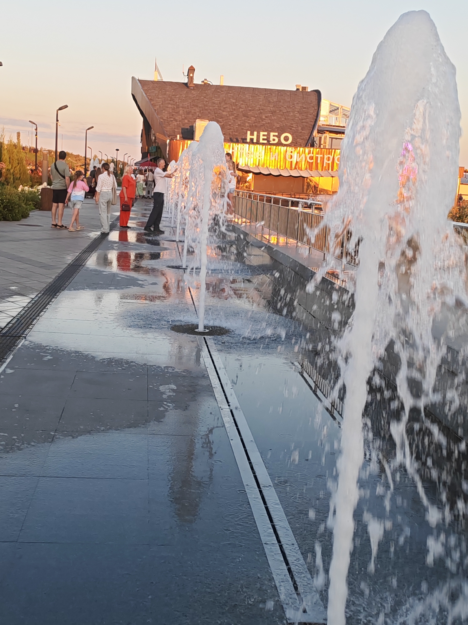 This is a medium shot, captured at eye-level, of a pedestrian promenade with a water feature in Tolyatti, Russia. The scene is illuminated by the warm, late afternoon sun, casting long shadows and a golden glow. In the foreground, a water jet from a fountain sprays upwards and to the right, blurring slightly due to motion. The ground is wet and reflective from the water feature, which consists of a series of water jets emerging from evenly spaced channels. Several people are visible in the mid-ground, walking along the promenade. From left to right, a man with a dark t-shirt and shorts, a young girl in a pink top and shorts, a woman in a white top and light-colored pants, and a woman in an orange outfit are walking away from the camera. Further along, a man in a light-colored shirt and dark pants is seen interacting with the water feature. Behind the people and the water feature, a modern building with a dark, angled roof is prominent. The building features large windows and illuminated signage, with the word "НЕБО" (Sky) clearly visible in white letters on the upper facade, and "КФС и бистро" (KFC and bistro) visible below. The overall environment appears to be a well-maintained urban public space, likely a park or a waterfront area, designed for recreation and leisure. The sky is a clear, pale orange, indicative of sunset.