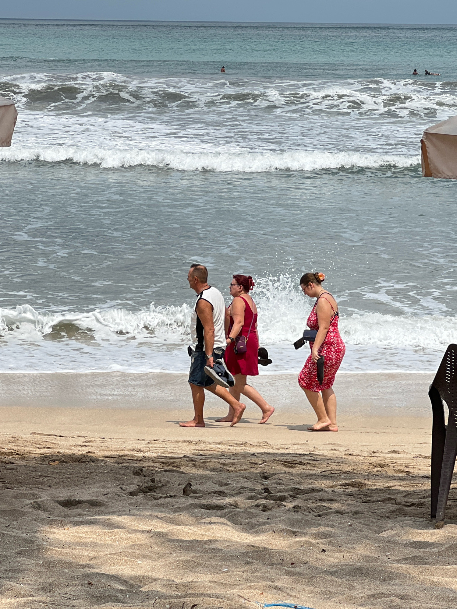 In Kuta, Indonesia, three individuals walk along a sandy beach adjacent to the ocean. A man in a white tank top and dark shorts walks on the left, carrying a pair of dark sandals in his right hand. To his right, a woman in a red dress with a dark bag slung across her chest walks, also barefoot. Further to the right, another woman in a red floral-print dress walks barefoot, holding a camera in her right hand. The ocean waves are breaking on the shore, and there are beach umbrellas visible in the background. The sand in the foreground is textured with shadows and a small bird.