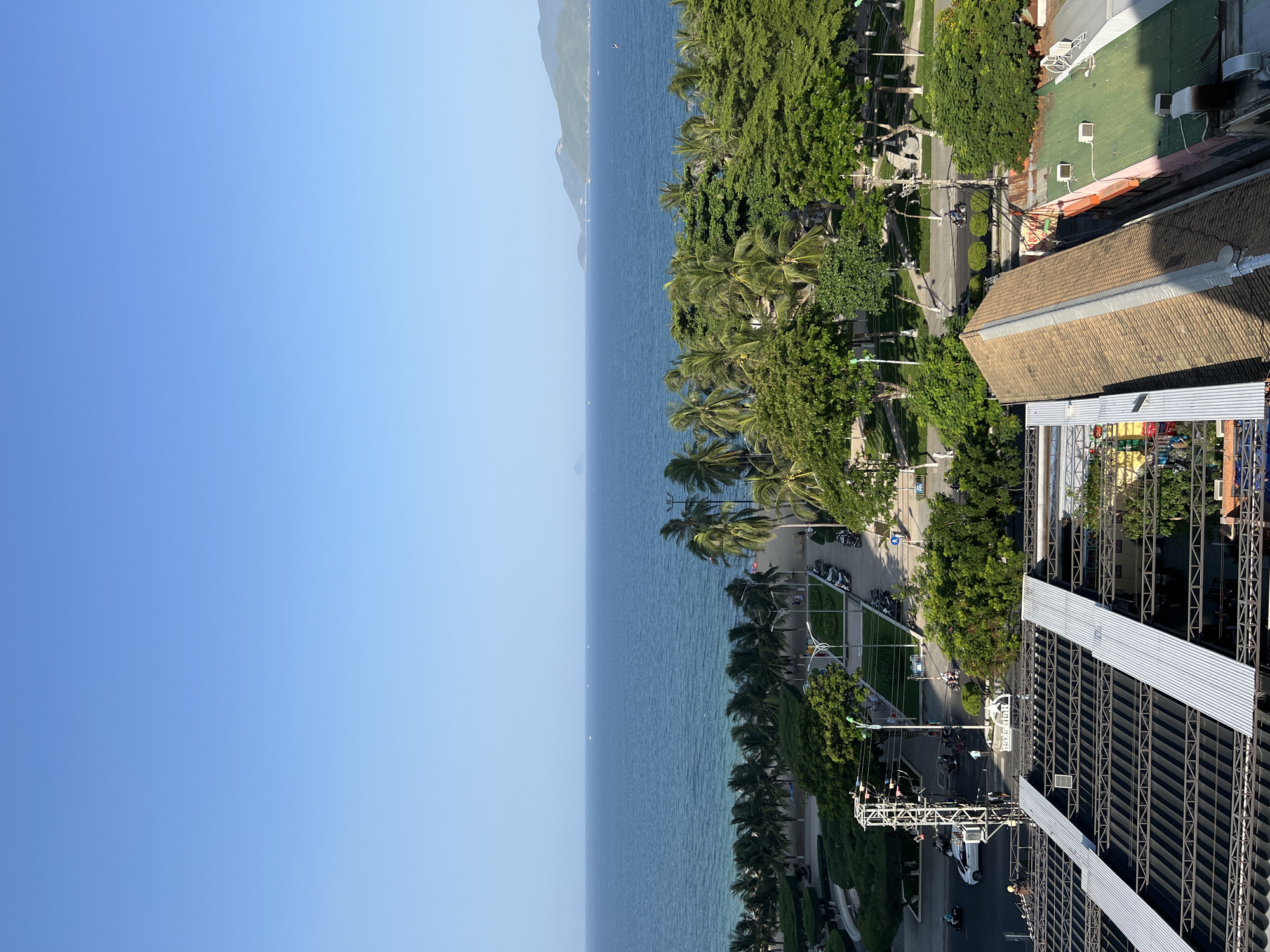 This aerial view captures a section of Nha Trang, Vietnam, featuring a coastal scene. The image is dominated by a clear blue sky and a vast expanse of blue ocean. Along the coastline, a row of palm trees separates the beach from a paved road lined with more trees and urban infrastructure. Several vehicles, including cars and motorcycles, are visible on the road, along with a few pedestrians. In the foreground, a section of a building's roof under construction, with metal beams and sheeting, is prominent. The overall environment suggests a sunny day in a developed coastal city.