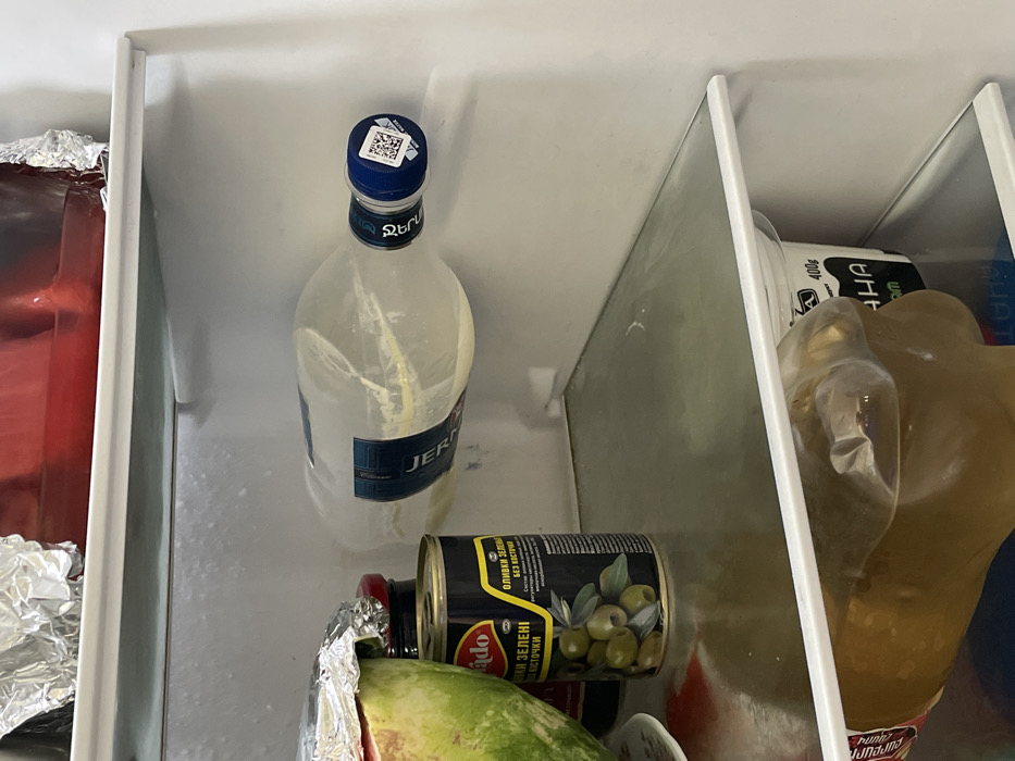 This image depicts the interior of a domestic refrigerator, located within a residence in Stepanavan, Armenia.
The scene shows three clear glass shelves containing various food items. The back wall of the refrigerator exhibits visible frost accumulation.
On the uppermost visible shelf, a red, rectangular food container is partially visible on the left, covered with crinkled aluminum foil.
The middle shelf holds multiple items:
* To the far left, a half-cut watermelon, with its green and white rind visible, rests on a white plate and is partially covered by aluminum foil.
* Next to the watermelon, a dark-colored glass jar with a red lid is present.
* Adjacent to the jar, a black metal can with a yellow label depicts green olives and is marked with Cyrillic text "ОЛИВКИ ЗЕЛЕНИ БЕЗ КОСТОЧКИ" (Pitted Green Olives).
* To the right, a clear plastic bottle, labeled "JERMUK," lies horizontally. The bottle contains a light yellow, opaque substance, indicative of it being frozen or partially frozen, with visible expansion of its contents. The bottle is sealed with a blue cap featuring a white QR code sticker.
The lowermost visible shelf contains two items:
* On the left, a clear, irregularly shaped plastic bottle holds a brown liquid.
* On the right, a white plastic container, likely cylindrical, is partially visible with black Cyrillic text and "400g" printed on its label.
No individuals are present in the image. The items are static within the refrigerator.