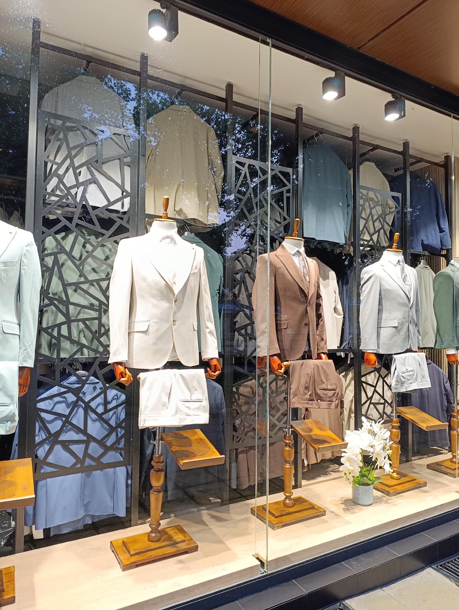 An exterior view of a men's clothing store window display shows several mannequins dressed in suits and shirts. The mannequins are arranged in rows with suits on display in a variety of colors including light blue, off-white, brown, and gray. Behind the mannequins, there are more suits and jackets hanging on racks, some with intricate geometric patterns on the wall behind them. The display is well-lit by spotlights. In the foreground, there are wooden stands holding suit pants. A small vase of white flowers sits on the floor of the display. The display is behind a glass window, with reflections of trees and the sky visible.