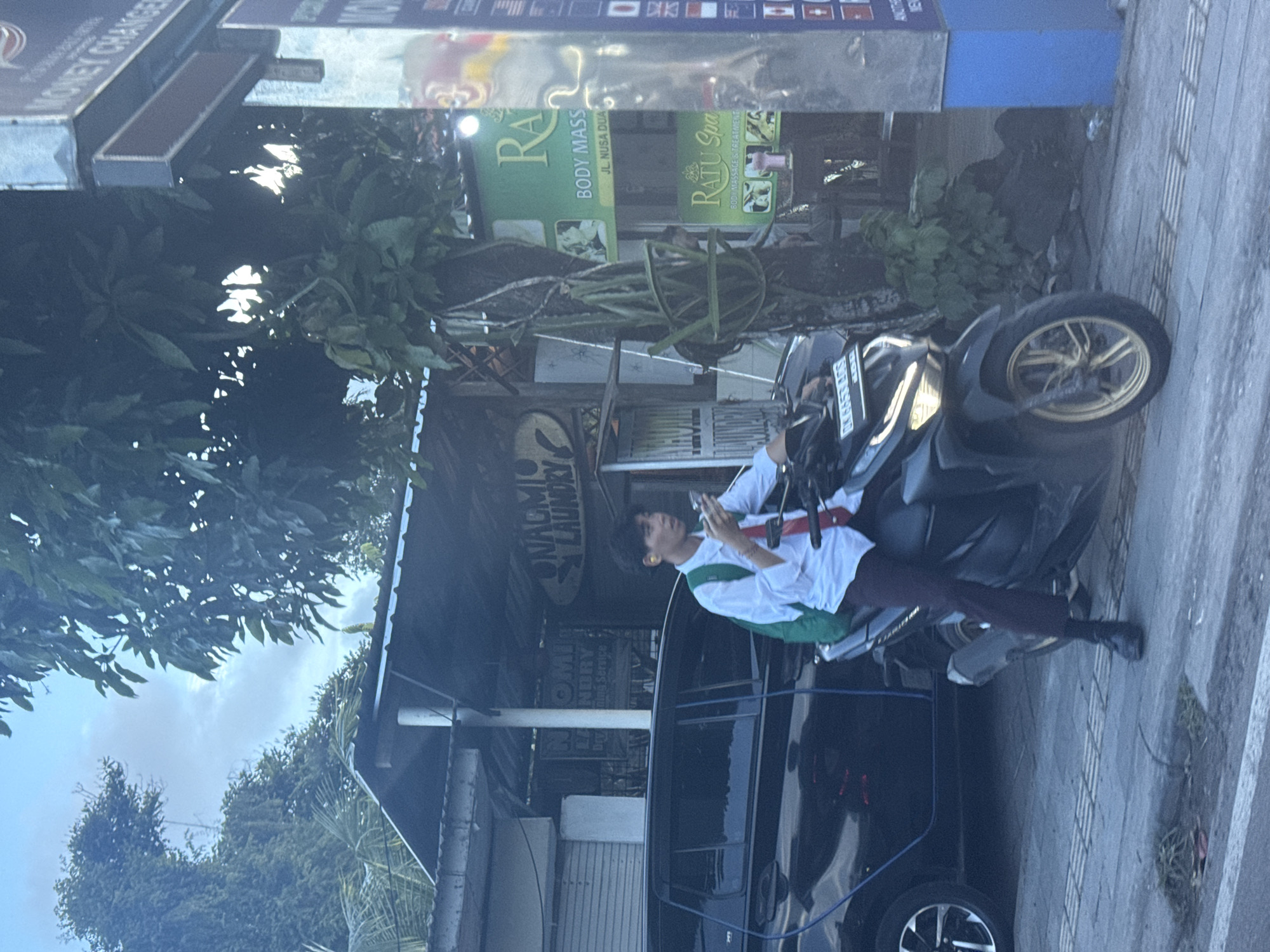 The image depicts a bright daytime street scene in Kuta Selatan, Indonesia, consistent with a commercial area, likely near Nusa Dua. A young person, wearing a white collared shirt, dark pants, and a green bag or strap, is seated on a black scooter with distinctive golden wheel rims. They appear to be engaged with their mobile phone. A black car is parked behind the scooter.
The street is flanked by various businesses. Prominently visible signs include "NALMIO LAUNDRY" which offers "Dry Cleaning Service", a "MONEY CHANGER", and wellness establishments named "RA BODY MASS" and "RATU Spa" both advertising "BODY MASSAGE & TREATMENT". The sign for "RA BODY MASS" specifically mentions "JL. NUSA DUA", confirming the location context. Lush green trees and foliage are present, suggesting a tropical environment, under a bright sky with some cloud cover. The license plate on the scooter reads "DK 6453 BES", further indicating a Bali location. No other people or significant traffic are visible, giving the scene a calm, unhurried atmosphere.