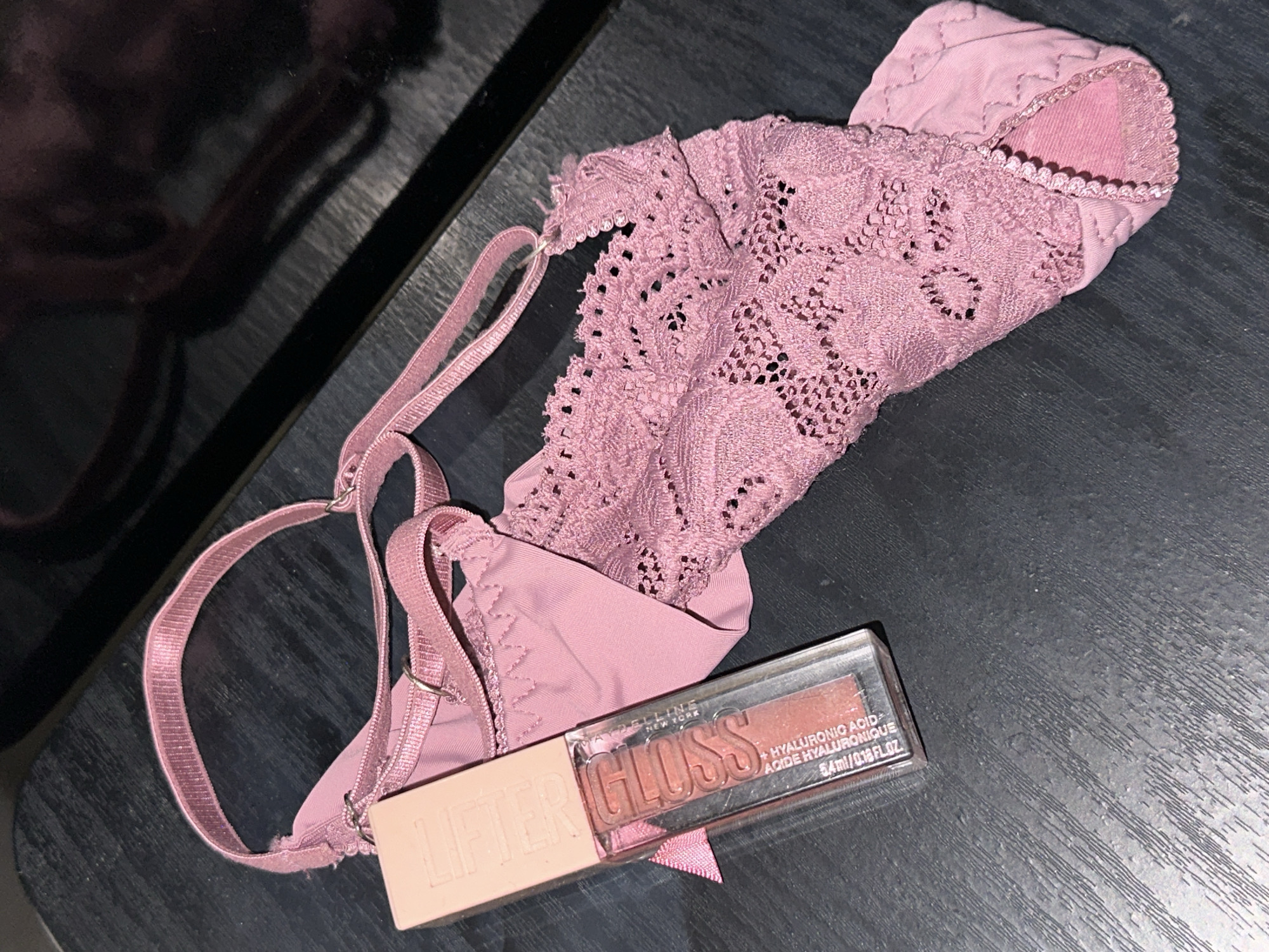 The image displays two items, a cosmetic product and an article of clothing, resting on a dark, textured surface. The surface appears to be a dark wood grain or laminate, exhibiting subtle linear patterns and variations in tone. In the upper right background, a dark, highly reflective surface is visible, possibly a polished panel or glass.
The primary object in the foreground is a clear rectangular tube of lip gloss. The brand "MAYBELLINE NEW YORK" is visible, along with the product name "LIFTER GLOSS" embossed on the light peach-colored cap. Text on the tube indicates "+ HYALURONIC ACID" and "ACIDE HYALURONIQUE," with a volume of "5.4 ml / 0.18 FL. OZ." The lip gloss itself has a shimmery, rosy-peach hue. A small, narrow pink ribbon is tied around the base of the applicator wand, visible within the tube.
Adjacent to and partially underneath the lip gloss is a pink, lacy garment, consistent with a bralette or delicate top. It features an elastic strap of matching pink, which is textured with fine horizontal ridges. This strap connects to the main body of the garment via small, silver-toned metal rings. The garment's main body is composed of intricate lace with a floral or geometric pattern, displaying numerous small perforations. Below the lace, a solid pink fabric section is visible, possibly a lining or a different material, with a subtly ribbed or quilted appearance at the bottom edge. A scalloped trim finishes a visible edge of the solid pink fabric. The garment is casually arranged, showing some folds and creases.
