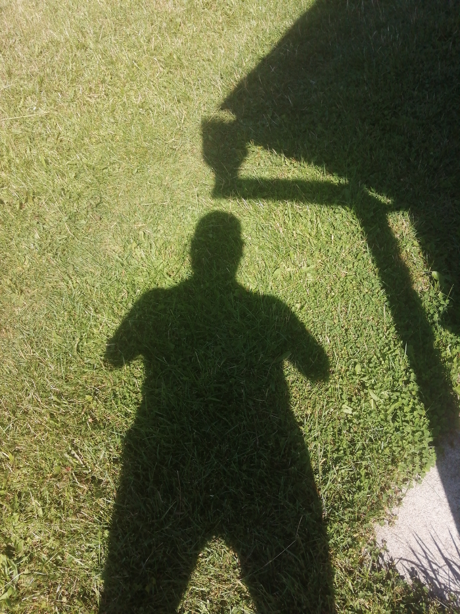 A human shadow is cast on a grassy lawn, with a portion of a building's shadow also visible. The lawn appears to be well-maintained, with short, green grass. The shadow of the person is in the center of the frame, appearing to be standing. The building's shadow is cast from the upper right corner of the image, suggesting it's a bright, sunny day. The location is identified as Slonim, Belarus.