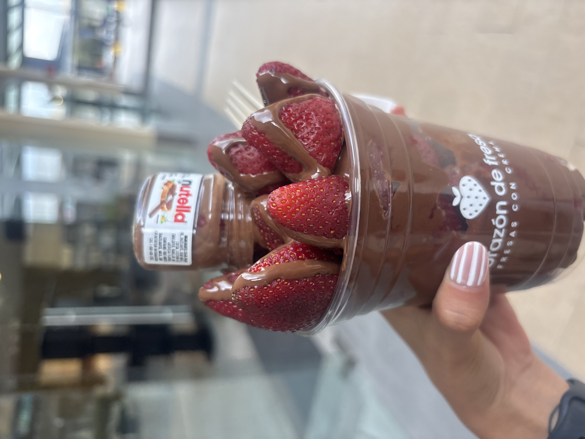 A close-up, sideways photograph showcases a hand holding a clear plastic cup filled with fresh strawberries and chocolate. The cup contains numerous bright red strawberries, heavily drizzled and coated with a thick, dark brown chocolate sauce, likely Nutella. A small, inverted Nutella jar is embedded within the cup, with its opening facing downwards, suggesting it is continuously dispensing its hazelnut spread onto the strawberries. A white, slender utensil, possibly a fork, is partially visible among the fruit.
The hand holding the cup has a prominent thumbnail adorned with a design of white and light brown vertical stripes. A black smartwatch or wristband is visible on the wrist of the person holding the dessert.
The plastic cup features branding: the words "corazón de fresas" (strawberry heart in Spanish) are prominently displayed in white text, accompanied by a stylized white strawberry logo. Partially visible text below reads "fresas con crema" (strawberries with cream) and a website address, "fresas.com". The small Nutella jar clearly shows the "nutella" brand name and some fine print detailing ingredients and contact information.
The background is softly blurred, indicating an indoor setting, likely a commercial establishment such as a mall, food court, or a specialty dessert shop in Puebla, Mexico, where this type of snack would be sold. The image captures the dessert as if it's just been prepared or is about to be enjoyed.
