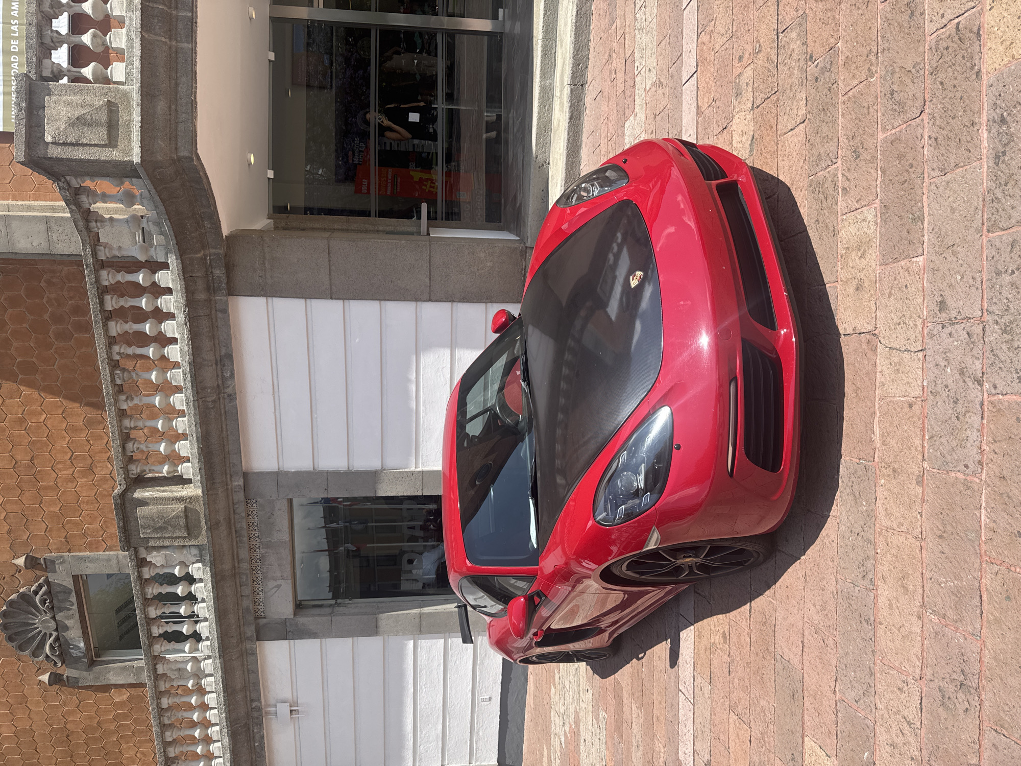 A red Porsche 718 Cayman GT4 sits on the brick sidewalk of Avenida 2 Norte in San Andrés Cholula, Mexico. The car is parked perpendicular to the façade of the Universidad de las Américas Puebla (UDLAP). The building has a classical architecture style with white columns and arched windows. A large glass door with an internal advertisement is visible behind the car. The day is sunny.