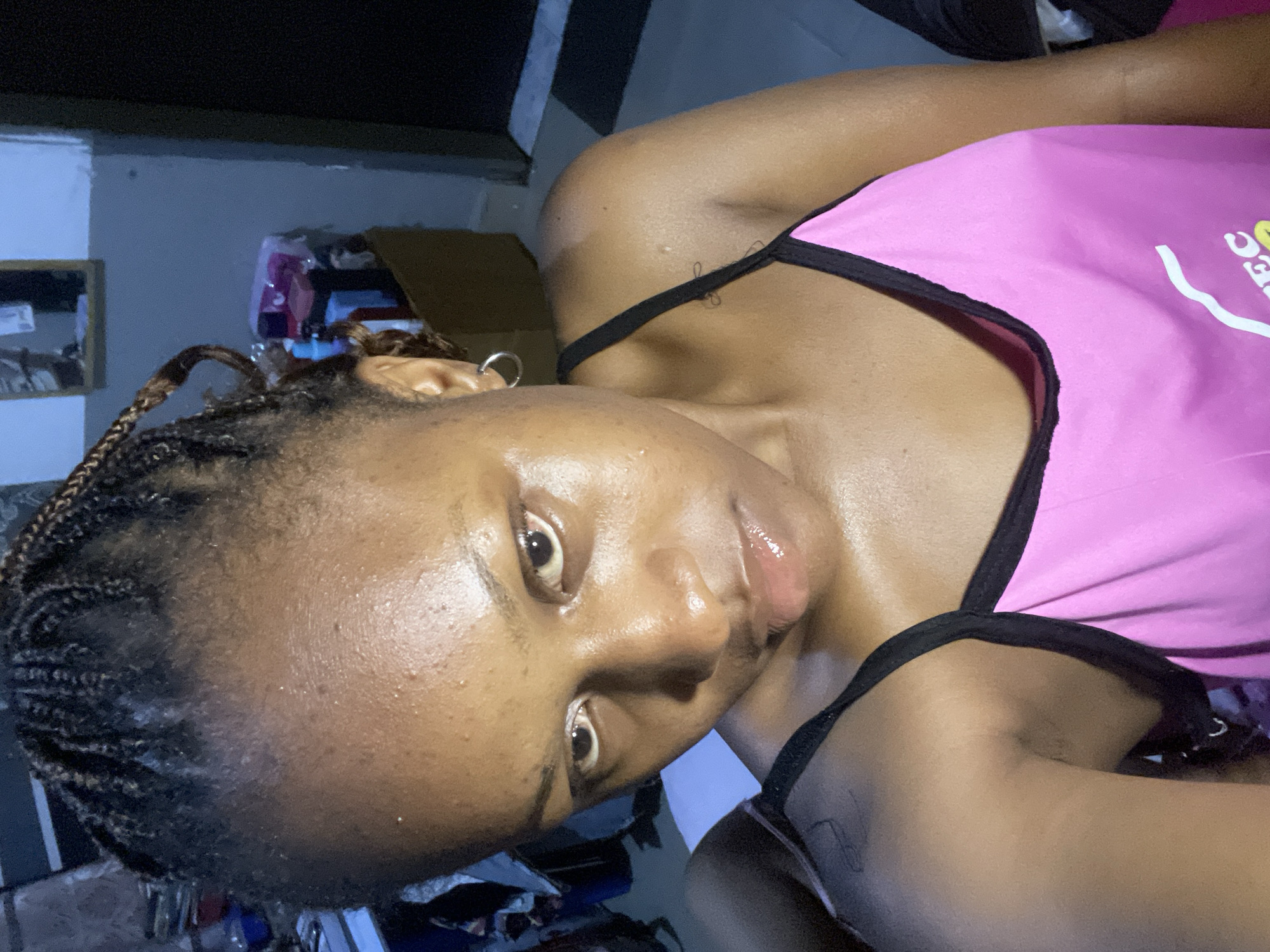 This is a close-up, selfie-style image of a young Black woman with braided hair and clear skin. She is wearing a bright pink tank top with black trim. The background appears to be an indoor setting, possibly a room in Rafin Par, Nigeria, with shelves containing various items. The lighting is dim, casting a slight blue hue on the walls. The woman's expression is neutral as she looks directly at the camera.
