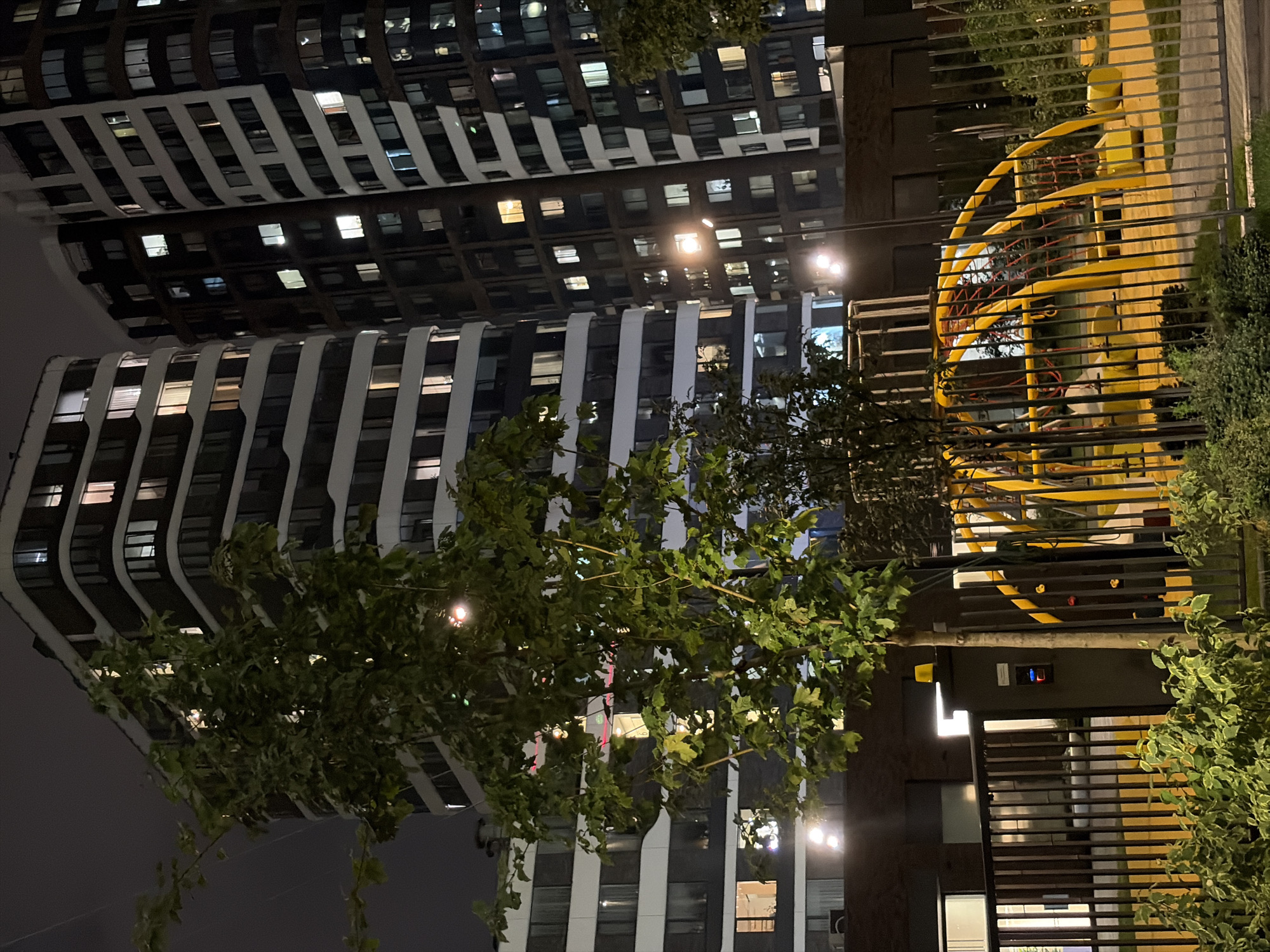The image depicts a nighttime view of two modern, high-rise residential buildings with illuminated windows, situated in an urban setting. In the foreground, a chain-link fence encloses a playground area with yellow climbing structures. A tree with green foliage stands prominently to the left of the frame, partially obscuring the buildings and the playground. The lighting suggests it is late evening or night, with streetlights visible, contributing to a sense of an active, yet calm, urban environment. Based on the architectural style and context, this scene is consistent with residential developments found in Moscow, Russia.