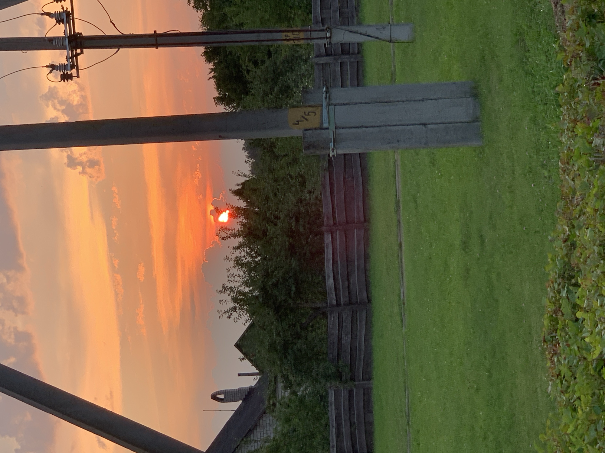 The image depicts a sunset scene in Kapyĺ, Belarus. The sky is filled with vibrant orange and pink hues, with clouds illuminated by the setting sun, which is partially obscured by clouds. In the foreground, there is a grassy field. Behind the field, a weathered wooden fence runs horizontally. Beyond the fence, several trees and some buildings are visible, including a house with a distinctive chimney. A power pole with electrical insulators and wires stands prominently in the right foreground. Another power pole is visible to the left. The overall atmosphere is tranquil and rural.