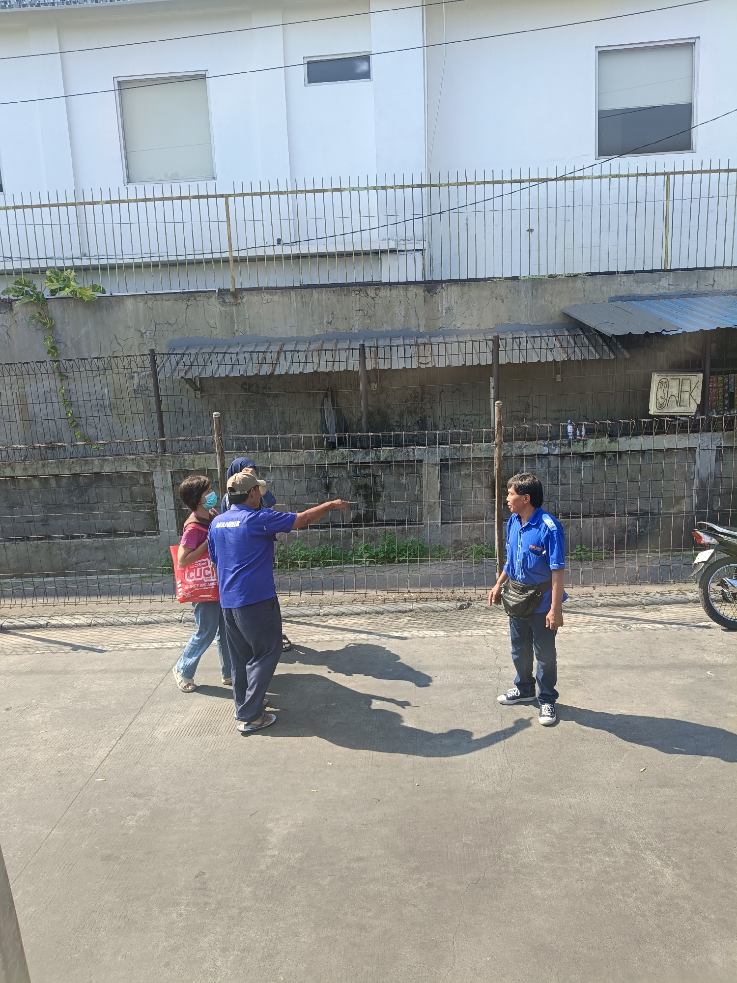 In Kedungrejo, Indonesia, a street scene unfolds under bright sunlight, casting sharp shadows. Three individuals are interacting on a paved walkway. A man in a blue shirt and dark pants, wearing a fanny pack, stands facing right, with his arm outstretched as if pointing or gesturing. To his left, a woman in a pink shirt and blue jeans, holding a red bag with Indonesian text, walks with a person in a headscarf. They appear to be engaged in a conversation or receiving directions. Behind them, a metal fence with vertical bars separates the walkway from a concrete wall, beyond which is a building with windows. A portion of a motorcycle is visible on the far right of the frame. The environment suggests an outdoor urban setting.