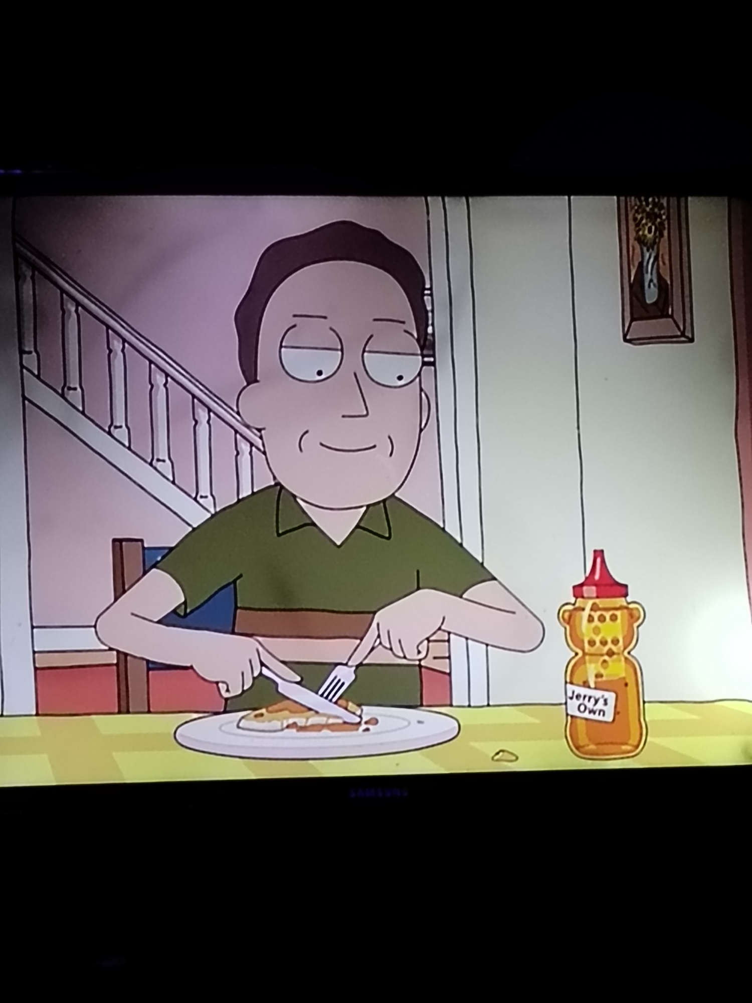 The image shows a cartoon character, likely Jerry Smith from Rick and Morty, sitting at a table and eating. He is wearing a green shirt with brown stripes and is using a knife and fork to cut into a piece of food on a white plate. To his right is a bottle of "Jerry's Own" honey, shaped like a bear. The background appears to be a dining room with a staircase and a framed picture on the wall. There is no information in the image that indicates a location in the city of Du, Nigeria.