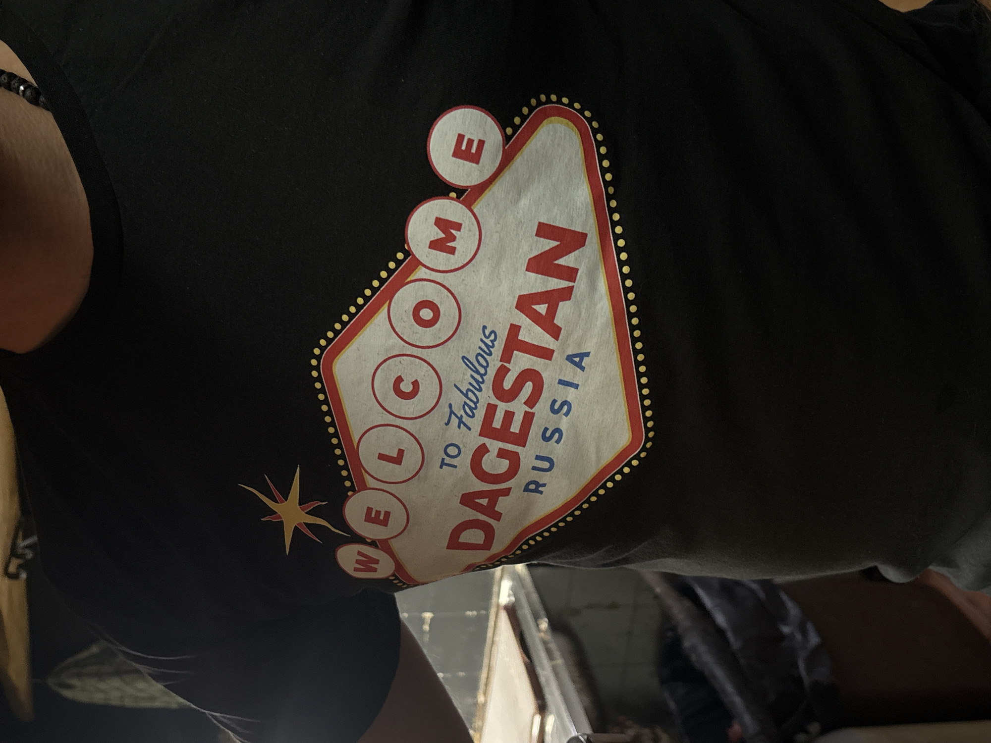 A close-up shot captures a person wearing a black short-sleeved t-shirt, with part of their torso and left arm visible. A dark, beaded bracelet adorns the left wrist. The t-shirt features a prominent graphic design, mimicking the iconic "Welcome to Fabulous Las Vegas" sign.
The graphic is shaped like a stylized shield with a red outer border and a white interior, encircled by small yellow dots resembling light bulbs. On the left side, the word "WELCOME" is spelled vertically, with each letter enclosed in a red-bordered circle. To the left of "WELCOME," there is a yellow and orange starburst design. Inside the main white area, the phrase "to Fabulous" is written in a cursive, light blue font, followed by the bold, red text "DAGESTAN RUSSIA."
The background is out of focus, showing blurry shapes and shadows that suggest an indoor setting. The lighting appears natural, indicative of daytime. While the t-shirt's design references Dagestan, Russia, the photograph was taken in Vrnjacka Banja Municipality, Serbia. The image focuses on the intricate details of the t-shirt's graphic.