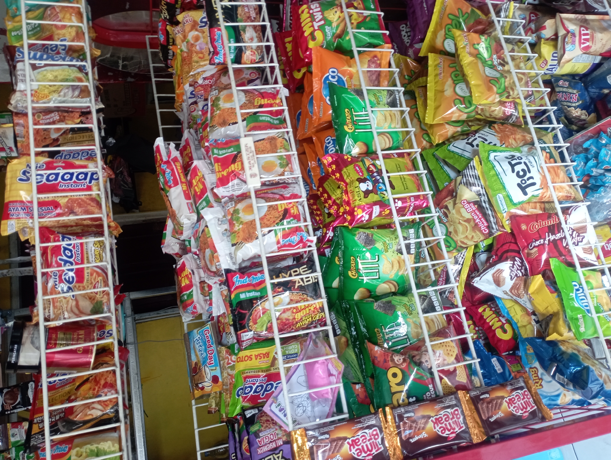 The image depicts a retail display of packaged food products, primarily instant noodles and snacks, arranged on white wire shelves. The products are diverse in their packaging colors and designs, with brands like "Mie Sedaap," "Indomie," "Chitato Lite," and "Tic Tac" clearly visible. Some packages feature images of food, while others have cartoon characters or text in Indonesian. The lighting suggests an indoor environment, possibly a small convenience store or warung. The overall impression is one of abundant, readily available packaged goods. The presence of Indonesian language on the packaging and the nature of the products strongly suggest the location is within Indonesia. Surakarta, also known as Solo, is a city in Central Java, Indonesia, and such displays are commonplace in its numerous small shops and markets. There are no people or specific identifiable landmarks present in the image, making it a generalized representation of a retail food display in Surakarta.