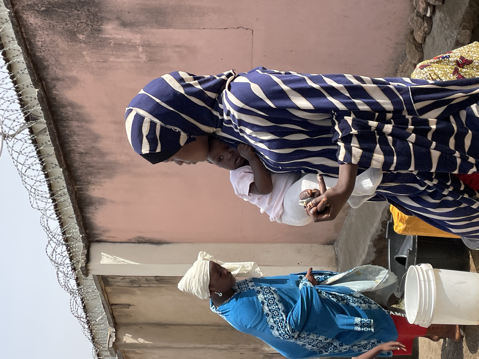 The image depicts two African women, one holding a child. The first woman is wearing a blue and white striped headscarf and dress, carrying a young child in a white shirt and light-colored pants. She is holding something in her hands. The second woman wears a blue dress with white trim and a white headscarf. She appears to be holding a white bowl or basin, and there are also white buckets nearby. The setting seems to be outdoors, possibly a courtyard or area near a building with a peach-colored wall and razor wire on the roof's edge. Based on the prompt, the location is Jos, Nigeria.