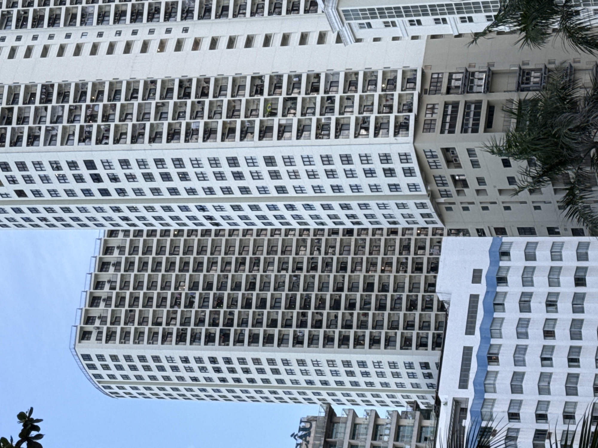 This is an aerial shot of multiple modern buildings under a clear blue sky, possibly taken from a drone or high vantage point. The buildings are predominantly white with repeating patterns of windows and balconies, suggesting they are residential or hotel structures. The architecture is characterized by sharp, clean lines and a grid-like facade, with balconies recessed into the structure. Some buildings appear to be connected or very close to each other, forming a dense urban landscape.
In the foreground and partially obscuring some of the buildings are lush green palm fronds, indicating a tropical or subtropical location. The light suggests it's daytime, with the sun casting shadows that highlight the architectural details of the buildings. There are no people visible, and the scene is devoid of any specific activity, presenting a static architectural study.
The image's orientation is unusual, with the buildings appearing to jut out at an angle, possibly due to the shooting perspective. One building on the right side has a striking vertical blue wavy design element, breaking the monotony of the white facade. The bottom of the image shows the corner of another building, also appearing to be a modern high-rise with a grid of windows. The overall impression is one of a contemporary, well-developed urban area with a tropical ambiance. There is no discernible text in the image, apart from what appears to be random OCR-generated characters in the provided crops, which are not part of the actual image content. The location context is given as Nha Trang Ward, Vietnam, which aligns with the presence of palm trees and modern coastal architecture.