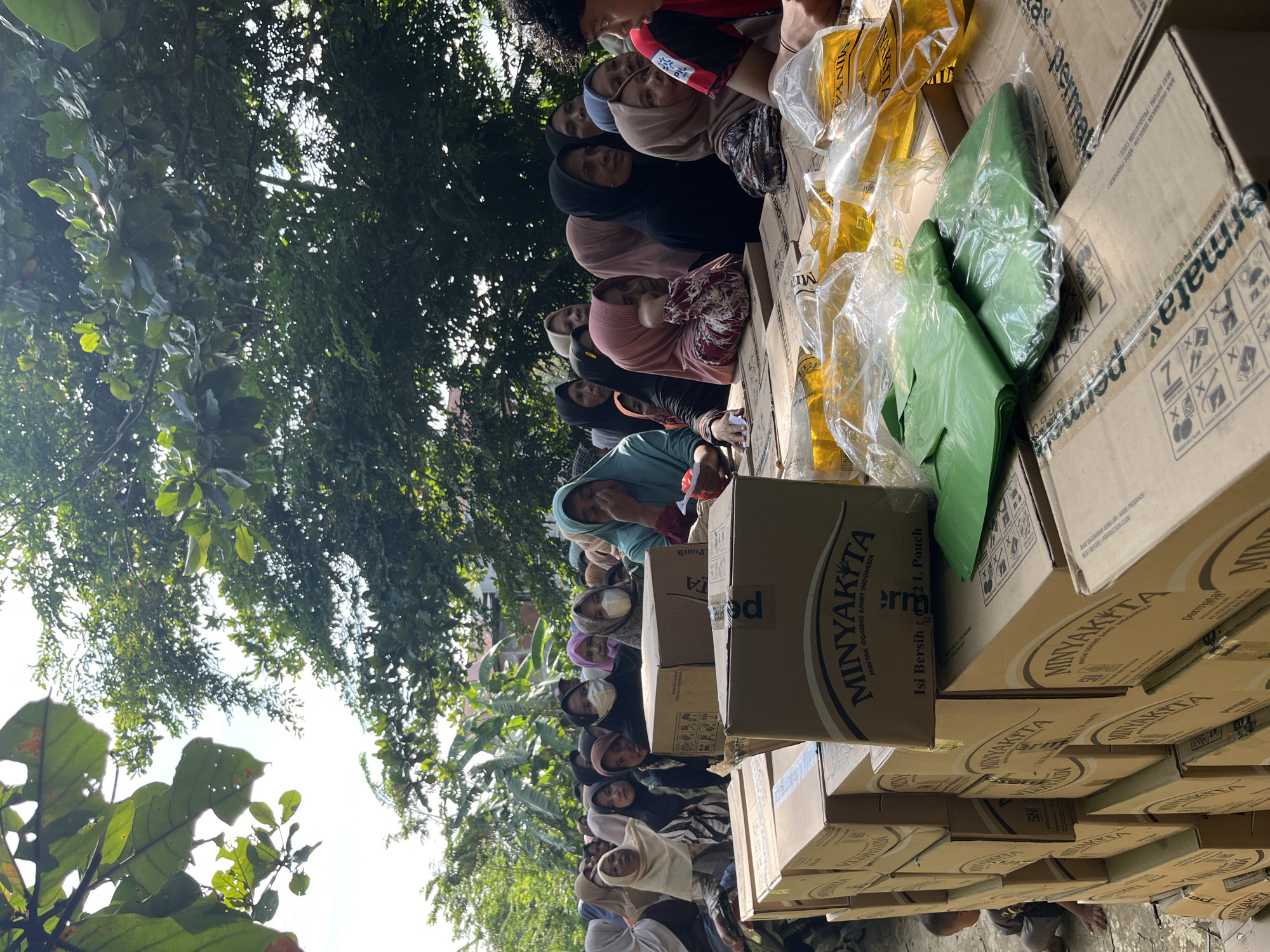 In Pulodolang, Indonesia, a group of people, mostly women wearing hijabs, are gathered outdoors, seemingly waiting in line. They are positioned behind stacks of cardboard boxes, many of which are labeled "MINYAKITA" and appear to contain cooking oil. Several pouches of cooking oil are visible on top of some of the boxes. The environment is partly shaded by trees, with the sky visible through the leaves. Some individuals are wearing face masks. The overall impression is of a distribution event, likely for food aid.