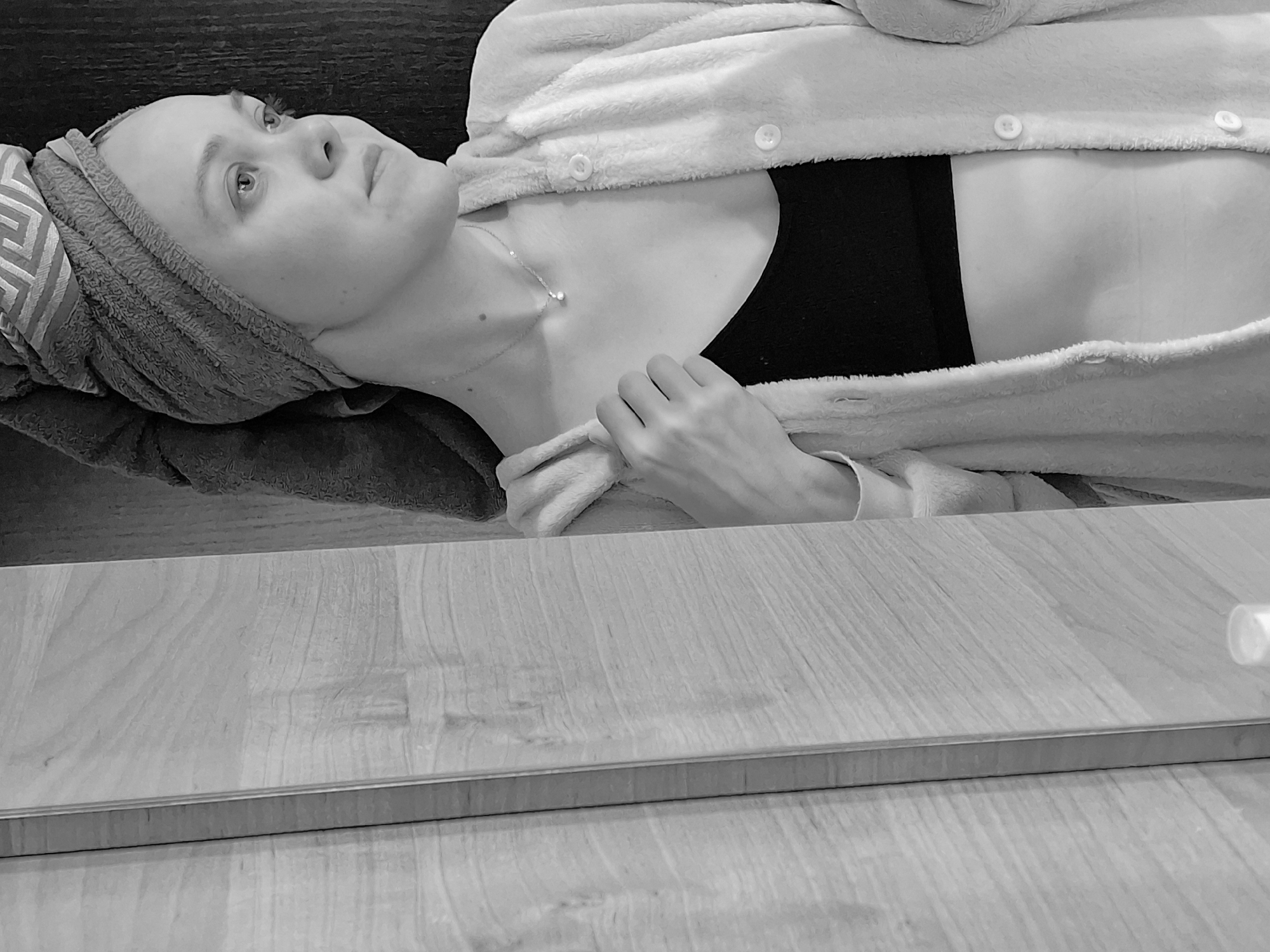 A pale-skinned woman is lying on a surface, her head resting on a dark-colored pillow or cushion. She is wearing a patterned towel wrapped around her head and a soft, plush robe that is open in the front. Underneath the robe, she is wearing a dark garment, possibly a top or bra. A delicate necklace adorns her neck. Her gaze is directed upwards and to the left. The background is dark and indistinct. In the foreground, there is a light-colored, wood-grain surface, possibly a table or counter. At the edge of the surface, a thin, metallic border is visible, and a small, white object sits near the edge. There is no indication about the location or information to relate the image to Chaikovsky, Russia.