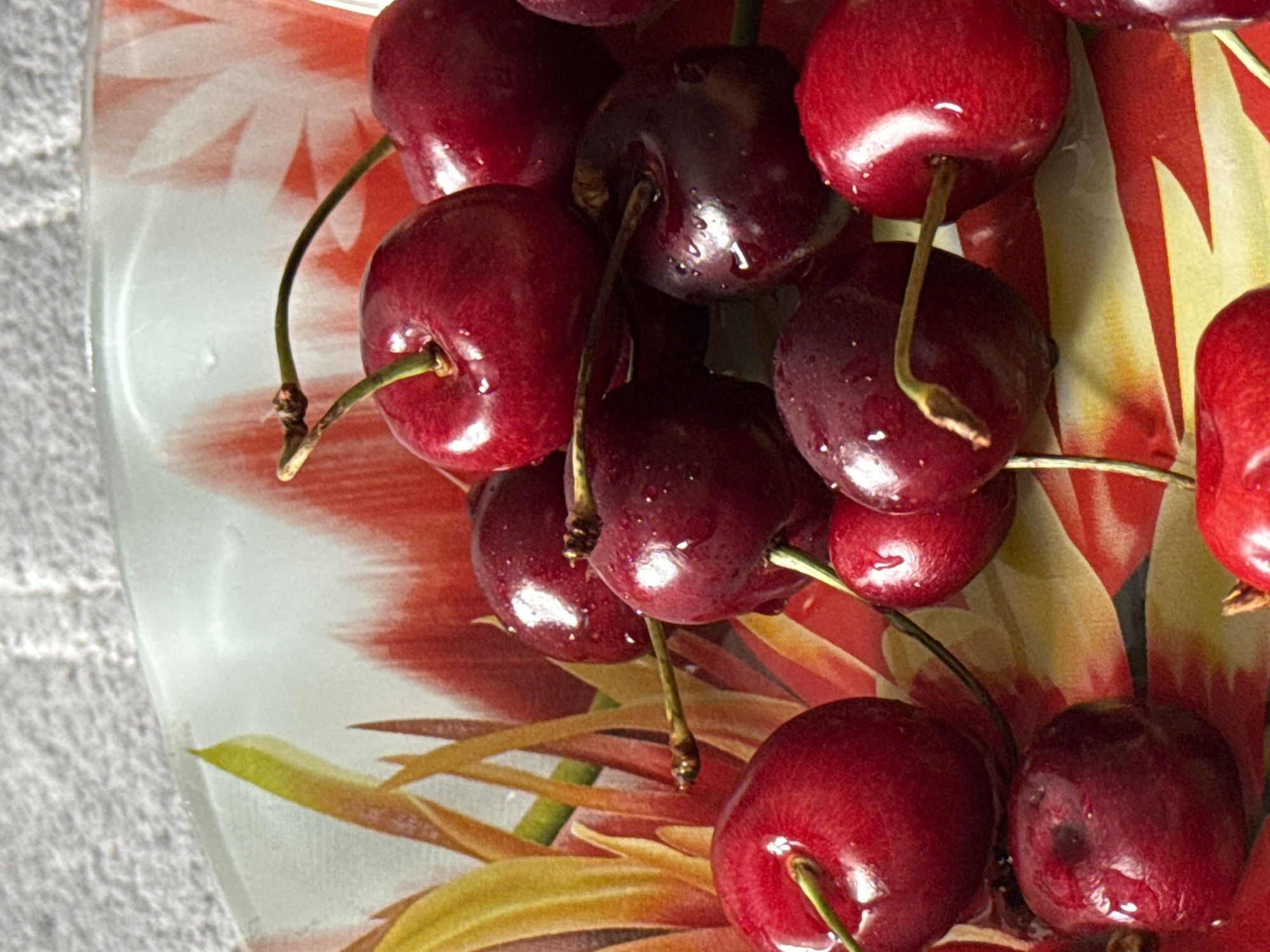 A close-up, overhead shot shows a pile of dark red cherries with water droplets on them, resting in a patterned bowl. The bowl features an abstract design of red and yellow brush strokes. The cherries are positioned in a somewhat haphazard cluster, with some stems visible. The lighting suggests it is daytime, with shadows cast by the cherries and the rim of the bowl. The background is a textured, off-white surface.