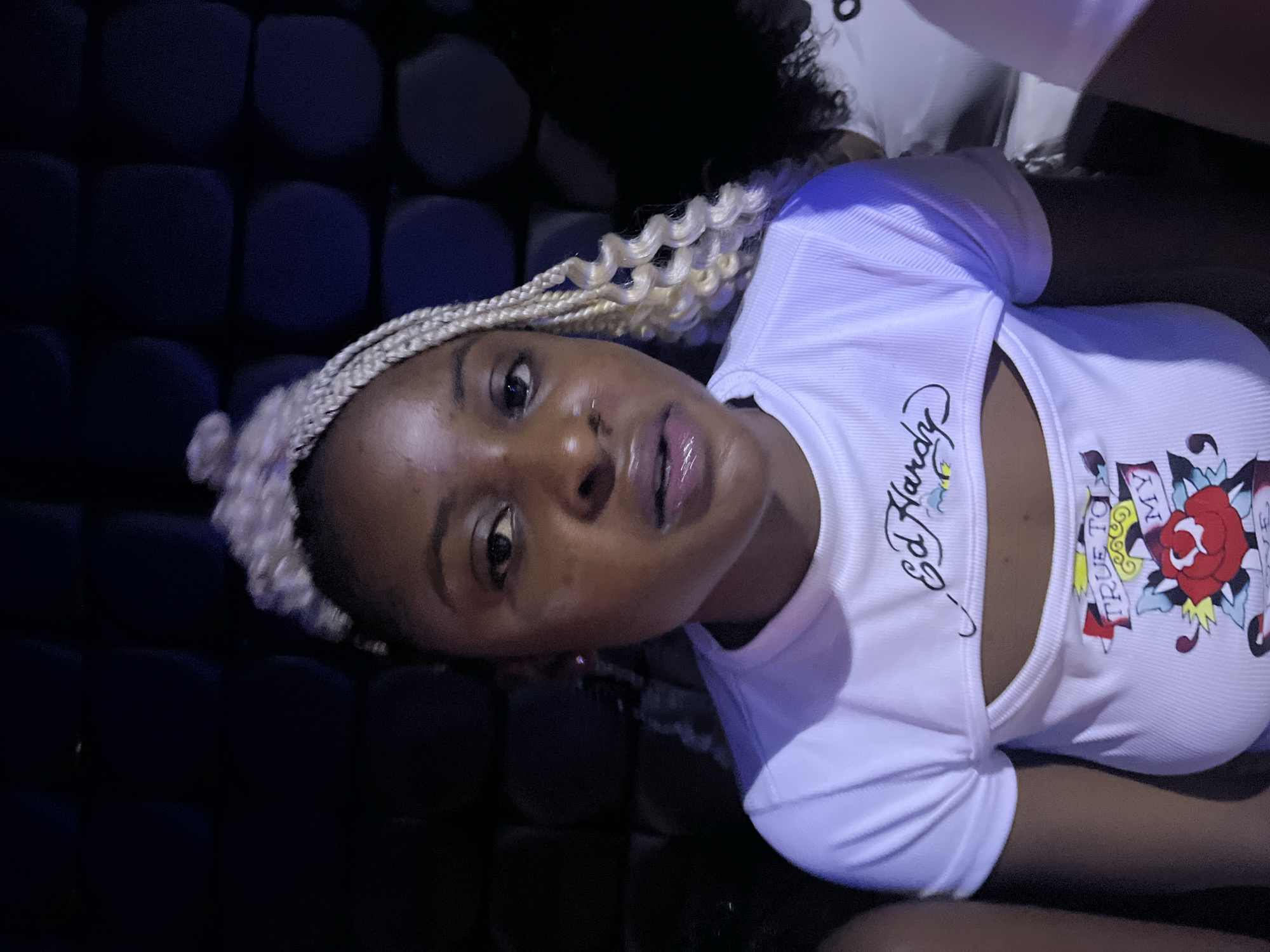 A young Black woman with blonde braided hair styled in a bun is the primary subject of the image, captured from the chest up. She is wearing a white, ribbed t-shirt with a cutout design on the chest. The shirt features graphic prints, including the "Ed Hardy" logo above the cutout and a rose tattoo design with the words "TRUE TO MY LOVE" below it. She has a nose ring and is looking directly at the camera with a slightly open mouth. The background consists of a dark, textured wall, possibly upholstered seating, creating a dimly lit atmosphere. The lighting suggests an indoor setting, possibly a club or entertainment venue, with some purple light casting onto the subject's clothing. The image appears to be a close-up portrait. There are hints of other individuals to the left and right, out of focus.