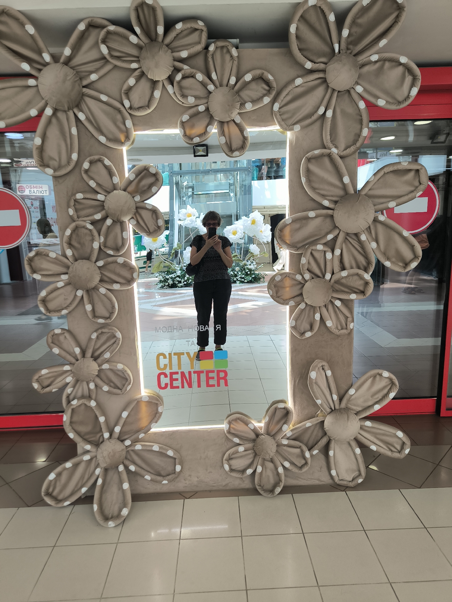 The image shows a large, flower-themed photo frame, likely made of plush material, positioned in a public indoor space. A woman is standing behind the frame, looking at her phone, and is reflected in the mirror of the frame. The frame is decorated with beige-colored flowers with white polka dots. Text on the frame reads "CITY CENTER". Based on the text "CITY CENTER" and the presence of the "City Center" shopping mall in Mykolaiv, Ukraine, it is highly probable that this image was taken at the "City Center" shopping mall in Mykolaiv. The background includes store entrances with red doors, reflections of other shoppers, and decorative white flowers.