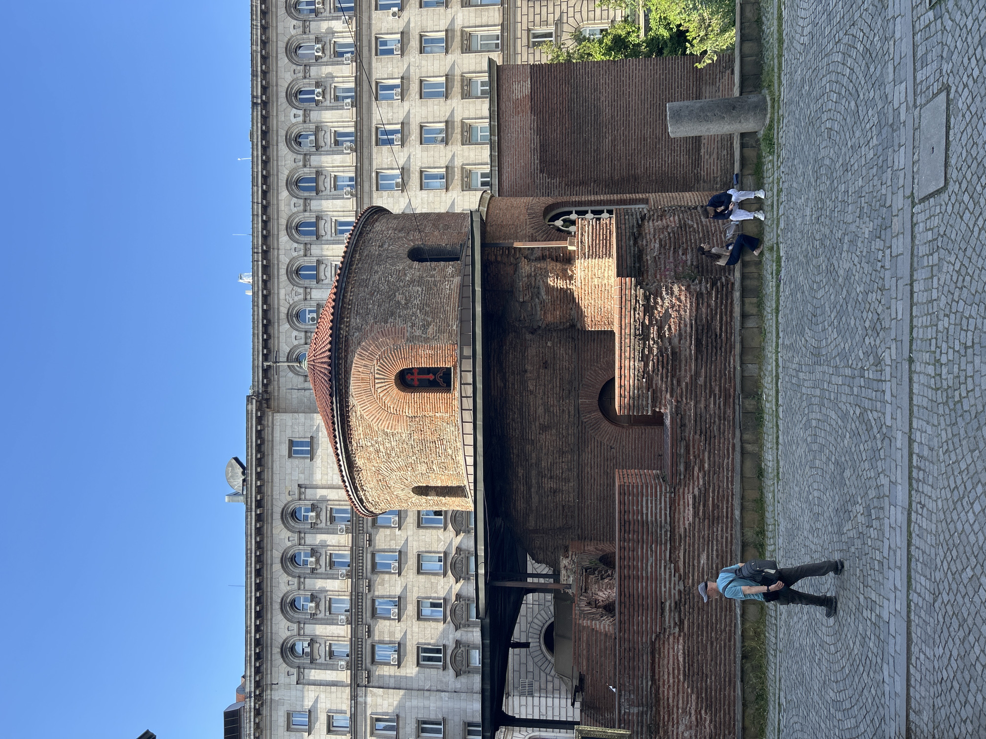 The image shows the Rotunda of Saint George in Sofia, Bulgaria. The rotunda, a red brick structure, is adjacent to a larger building with numerous windows and air conditioning units. A man in a blue shirt and gray pants is walking on a cobblestone path alongside the rotunda. Two other people are sitting on a raised ledge nearby. The sky is clear blue.