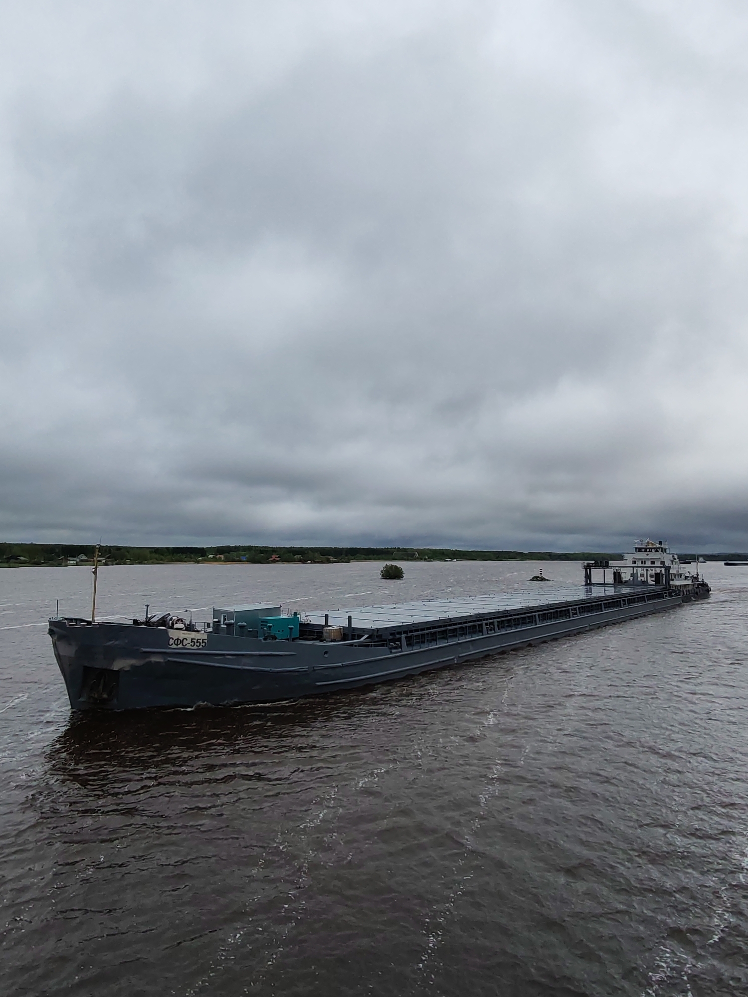 The image shows a large, gray barge moving through a body of water. The barge is long and rectangular with a superstructure at the rear and the Cyrillic letters "CФС-555" visible on the front. The water is choppy, indicating some wind. The sky is overcast with dense, gray clouds. A treeline is visible in the distance, and a small cluster of buildings is discernible among the trees on the left. This scene is in Shestovo, Russia.
