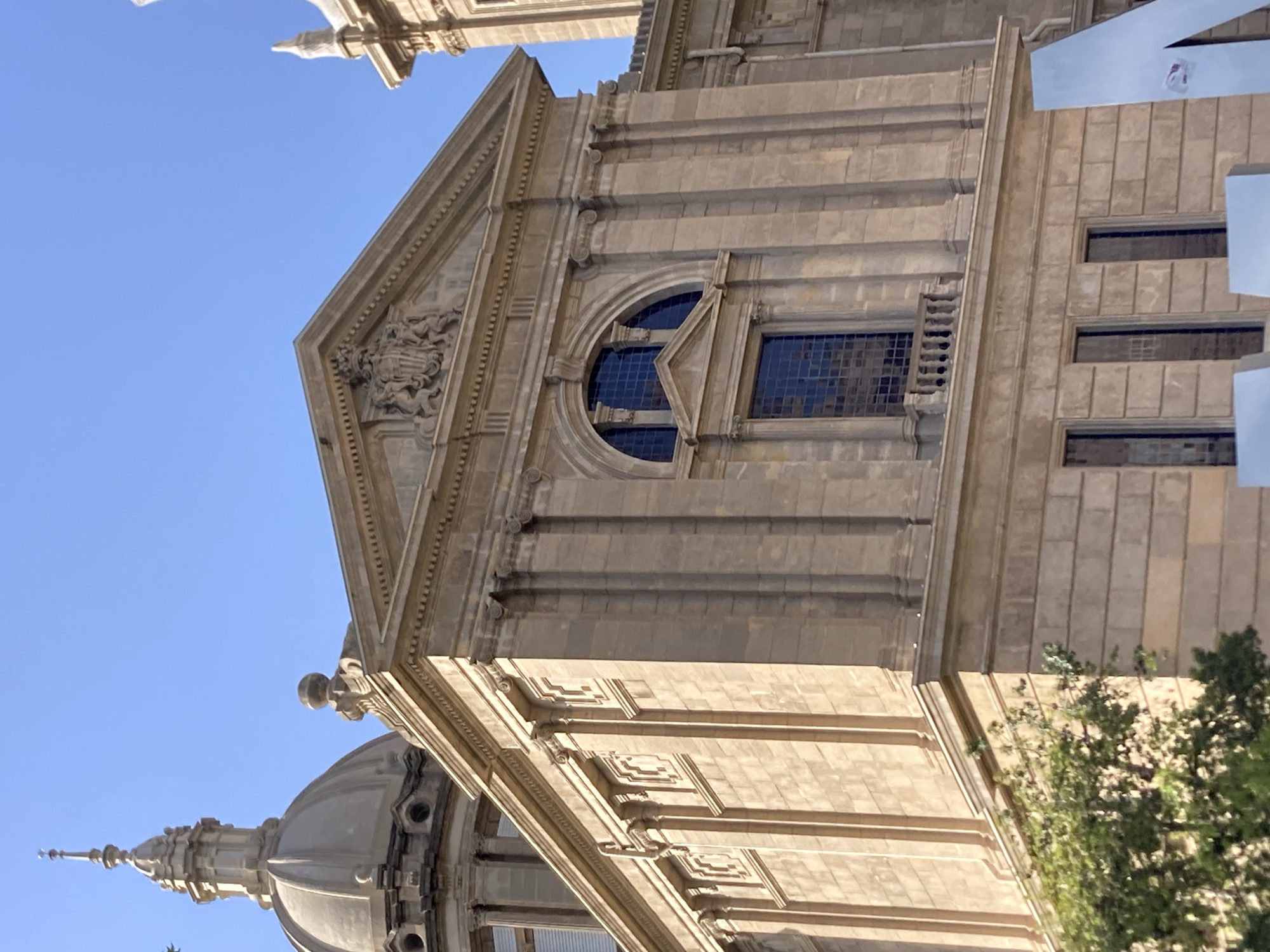 The image captures a close-up, low-angle view of the National Palace of Catalonia (Palau Nacional) in Barcelona, Spain, against a clear blue sky. The light beige stone facade of the building is highly detailed, showcasing classical architectural elements. Prominently featured is the central section of the building, with a triangular pediment at the top adorned with a coat of arms. Below the pediment is a large arched window with multiple panes, and directly underneath it, a smaller rectangular window with a balcony railing. The building's facade is decorated with vertical fluted columns and decorative moldings. To the left, a portion of a large dome with a spire is visible. In the foreground, the tops of green trees are partially obscuring the lower part of the building. In the bottom right corner, large, angled silver letters, possibly part of an art installation or signage, are visible. The lighting suggests it is daytime, likely mid-morning or afternoon given the bright sunlight casting shadows. There are no people visible in the frame.