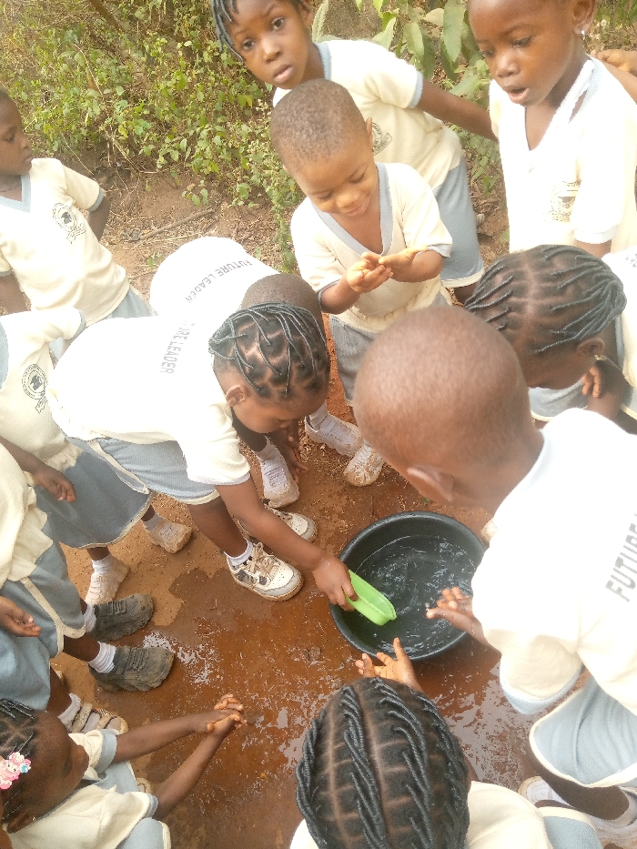 The image captures a group of approximately nine to ten young children in the city of Ukwu-Abwa, Nigeria. They are dressed in matching school uniforms consisting of cream-colored short-sleeved shirts, some featuring "FUTURE LEADER" printed on the back, and grey shorts or skirts. The children are gathered in a close circle outdoors on damp, reddish-brown earth, with mud visible on their footwear. At the center of the group, a black plastic basin filled with clear water rests on the ground. One girl, positioned center-left and wearing white and black sneakers, is actively stirring the water in the basin with a light green, leaf-shaped object. Another child, visible at the bottom of the frame, has hands clasped over the basin. Other children observe the activity, with various expressions of engagement and curiosity. The background reveals dense green vegetation and bushes.